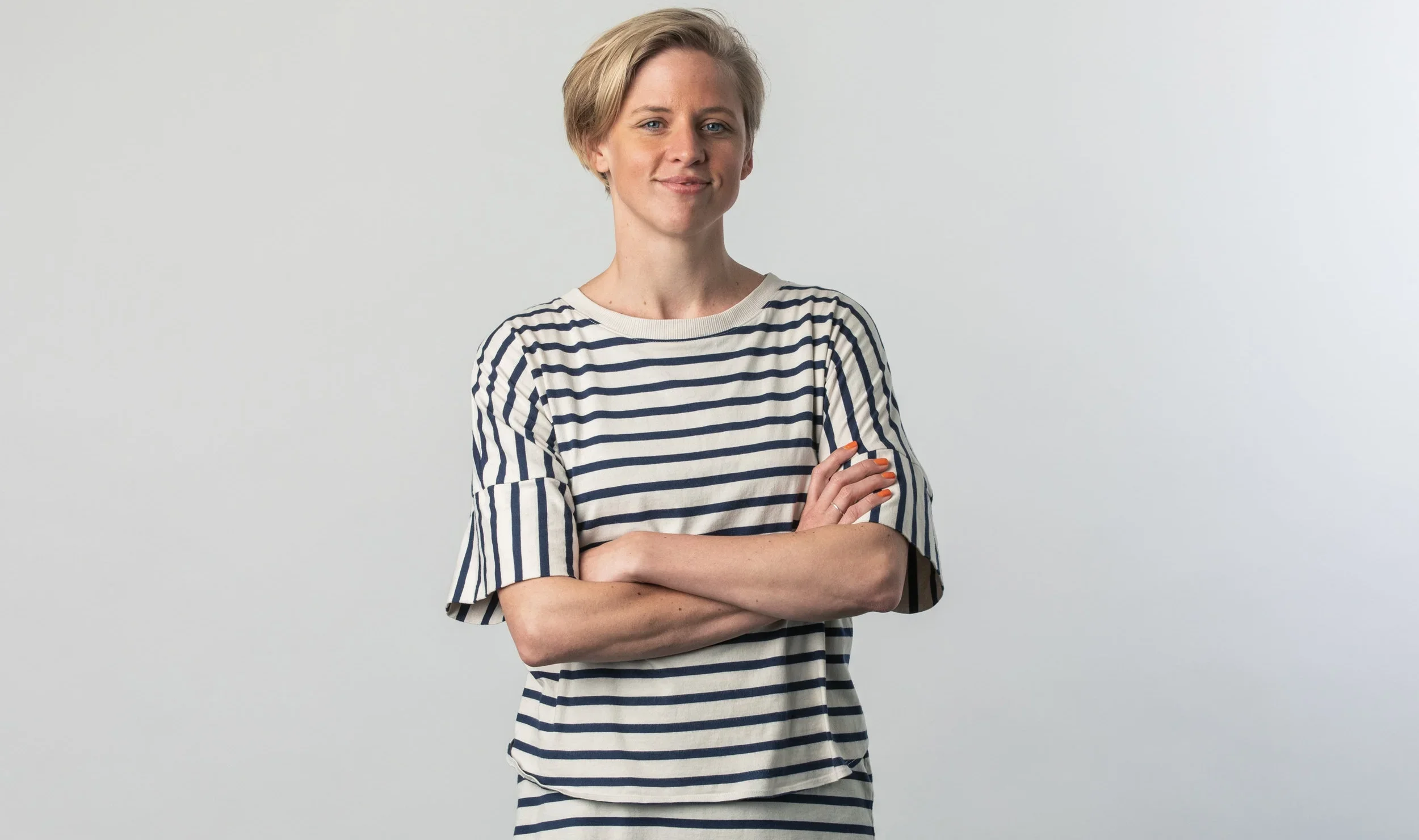 A woman with short blonde hair wearing a white and navy striped shirt, standing with arms crossed against a plain light gray background.