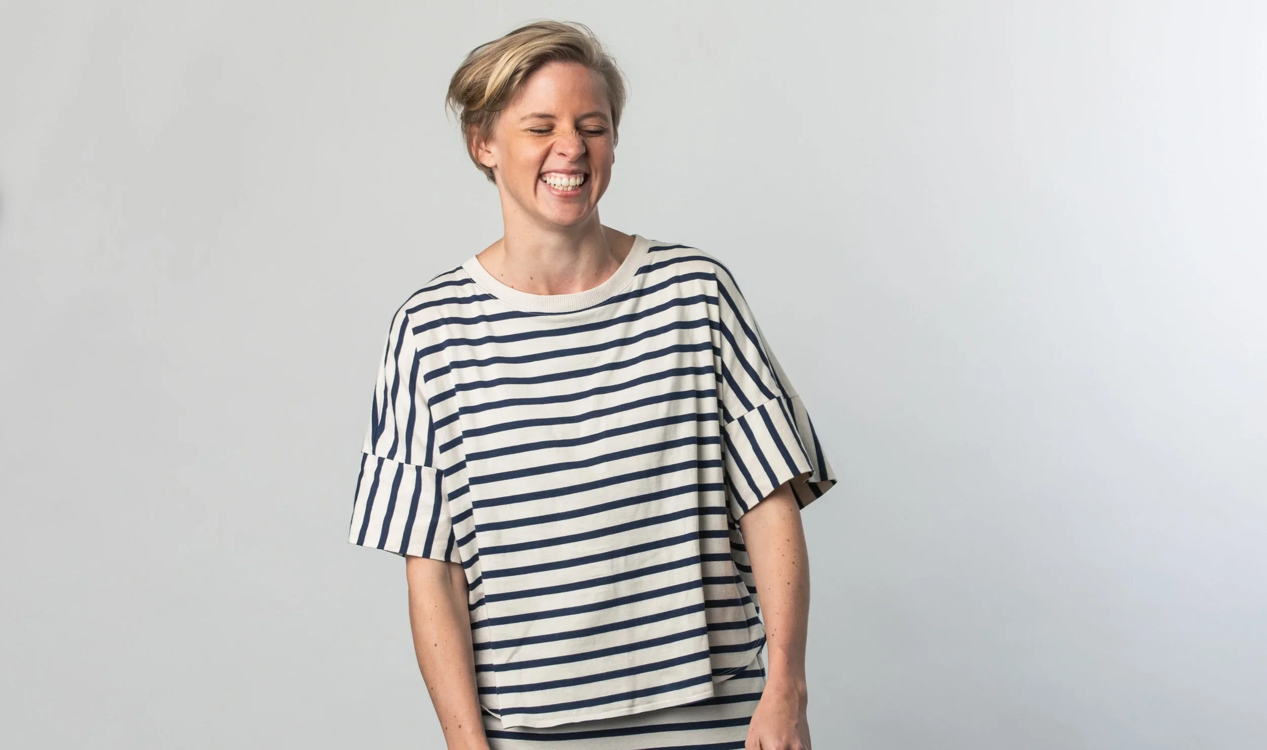 A woman with short blonde hair smiling and squinting, wearing a white and navy striped t-shirt, standing against a plain light gray background.