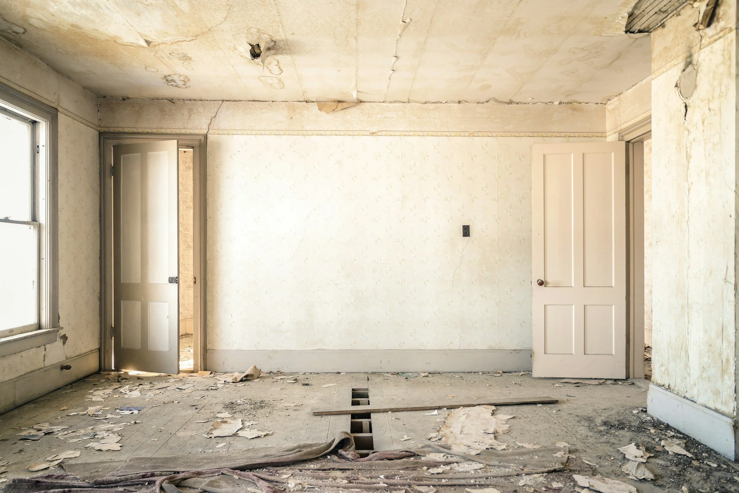 An empty, dilapidated room with peeling wallpaper, cracked ceiling, broken floorboards, and debris scattered across the floor.