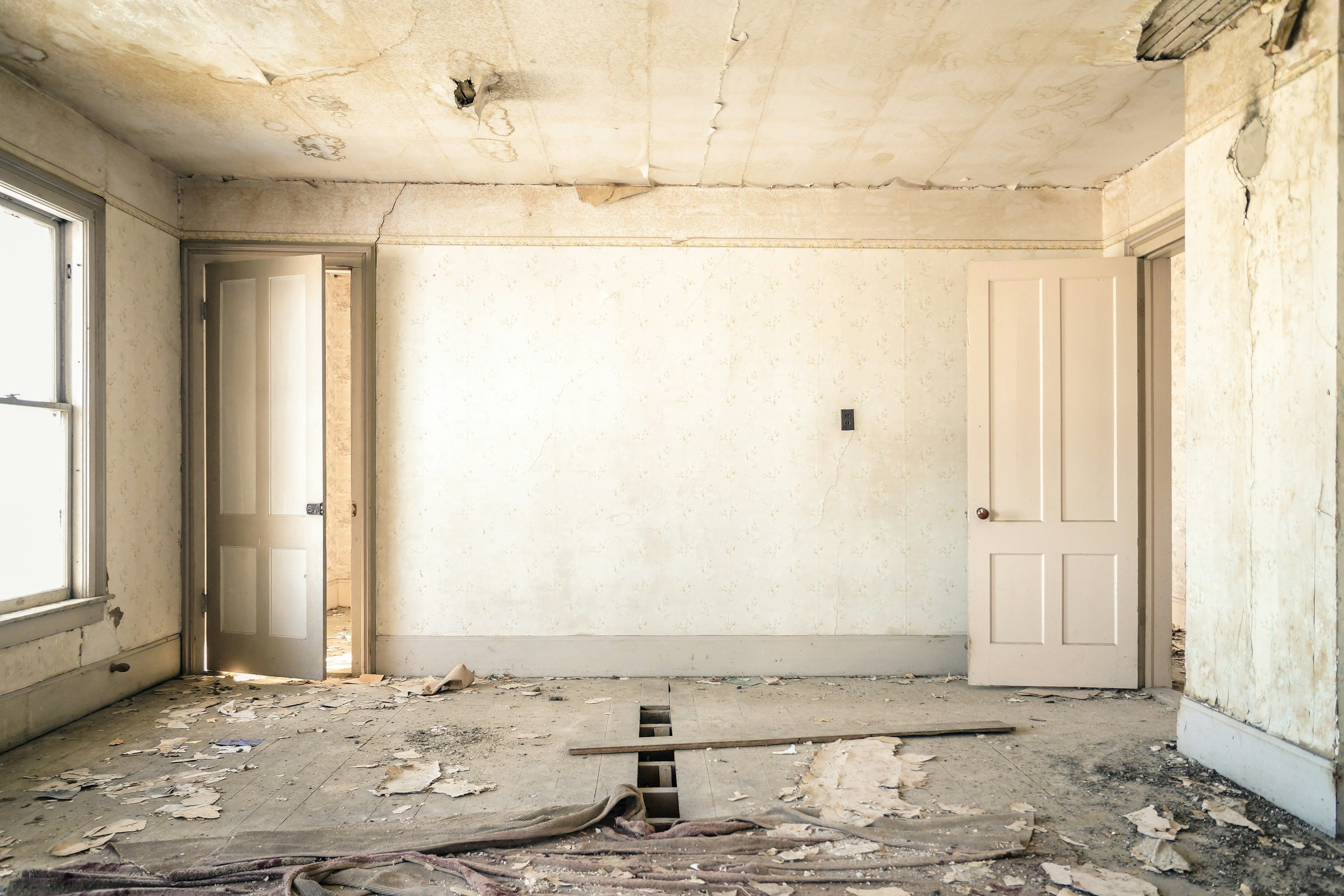 An interior room in disrepair with peeling paint and damaged drywall. The floor is covered in debris and peeling wallpaper, with holes in the ceiling. A window is on the left side, and a door is partially open on the left wall, leading to another room.