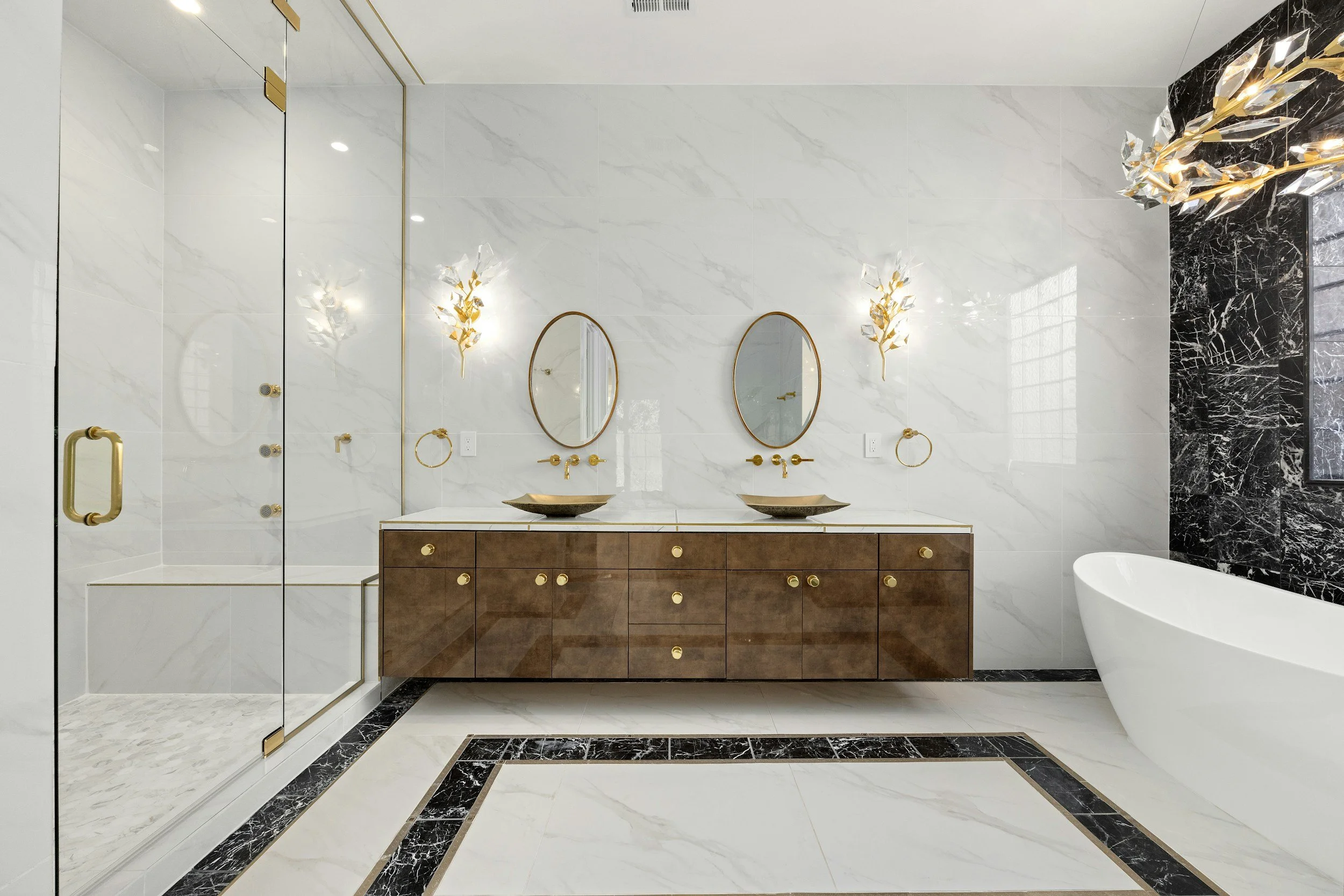 A modern bathroom with a marble wall and black marble accents. It features two oval mirrors above a wooden vanity with two vessel sinks and gold fixtures. There are decorative gold and glass wall sconces, a glass-enclosed shower with gold hardware, and a white freestanding bathtub.
