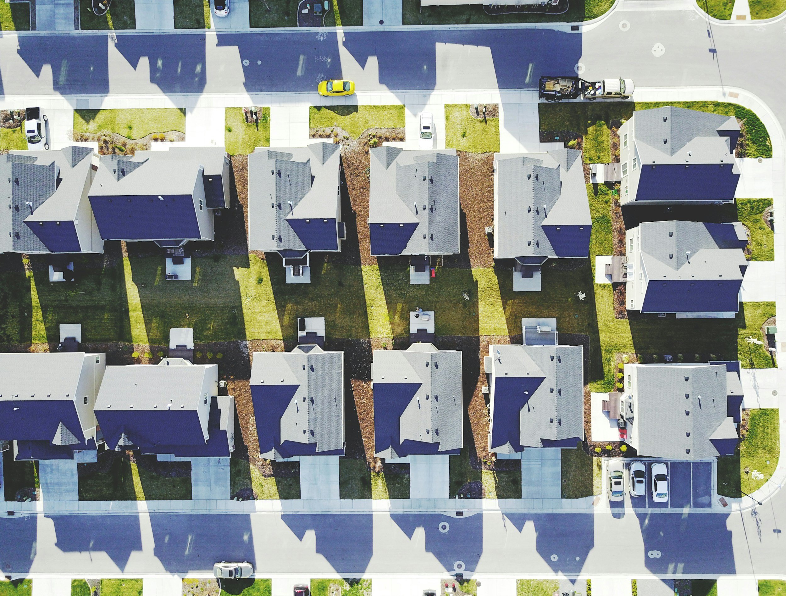 An aerial view of a residential neighborhood showing multiple houses with grey roofs, driveways, cars parked in the driveways and streets, green lawns, and sidewalks.
