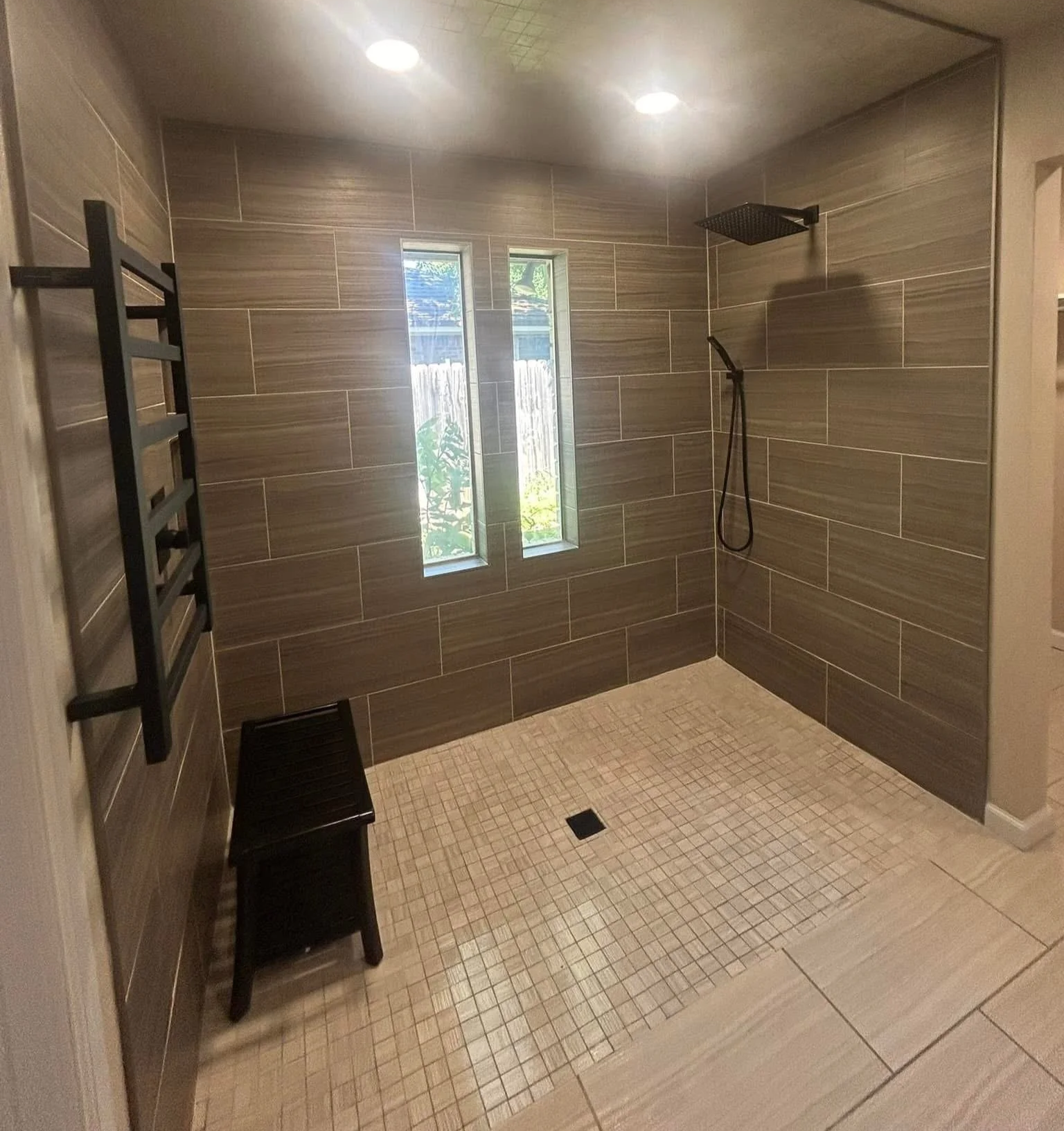 A modern walk-in shower with beige and brown tiled walls, a black ceiling-mounted rainfall showerhead, a hand-held shower, a small window, a black towel rack, a wooden stool, and a floor drain.