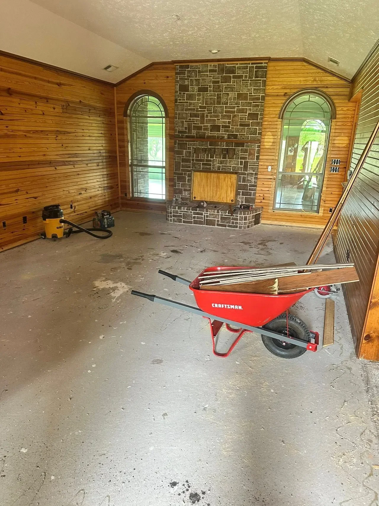 Construction or renovation room with wood-paneled walls, a stone fireplace, and large arched windows. There is a red wheelbarrow filled with wooden planks, a yellow shop vacuum, and a small portable power tool on the floor. The space appears under construction or renovation.