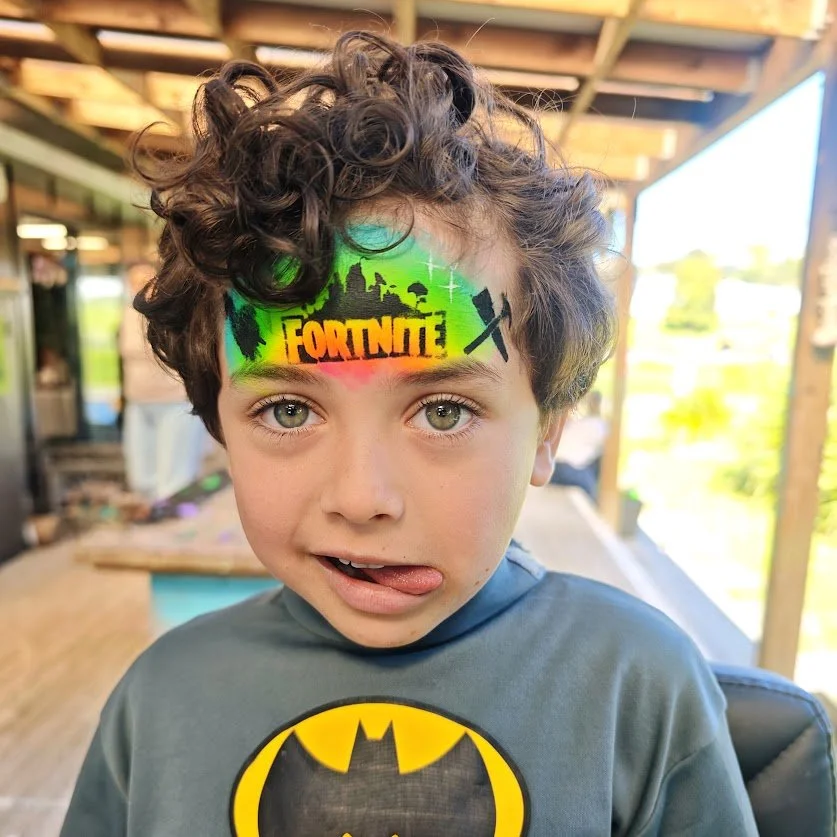 A young boy with curly brown hair and light-colored eyes making a playful face with his tongue out. He is wearing a gray shirt with a Batman logo. The boy has colorful face paint on his forehead with a Fortnite theme, including the game's logo, and s
