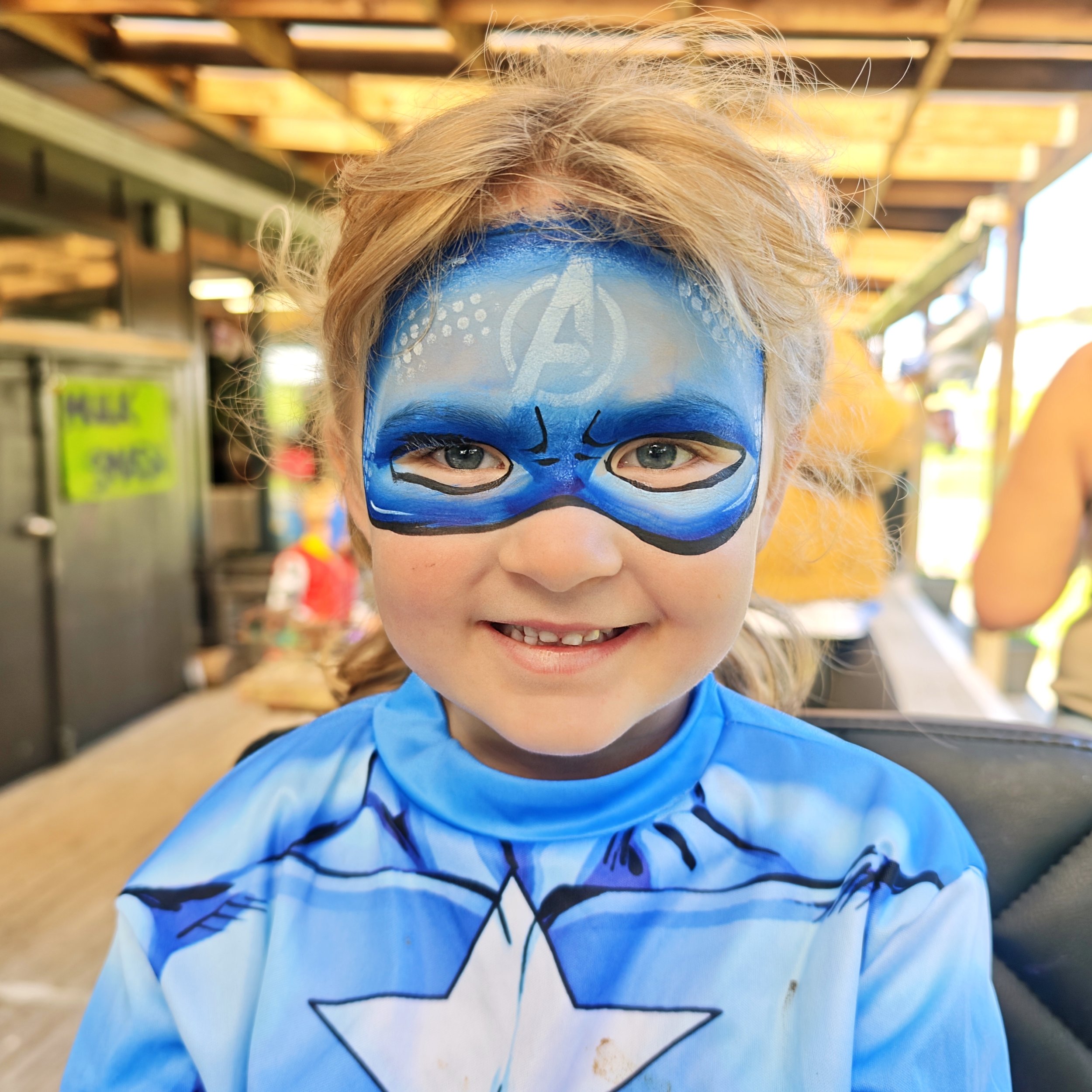 A young girl with face paint resembling a blue superhero mask, smiling outdoors with a blurred background of wooden structures and greenery.