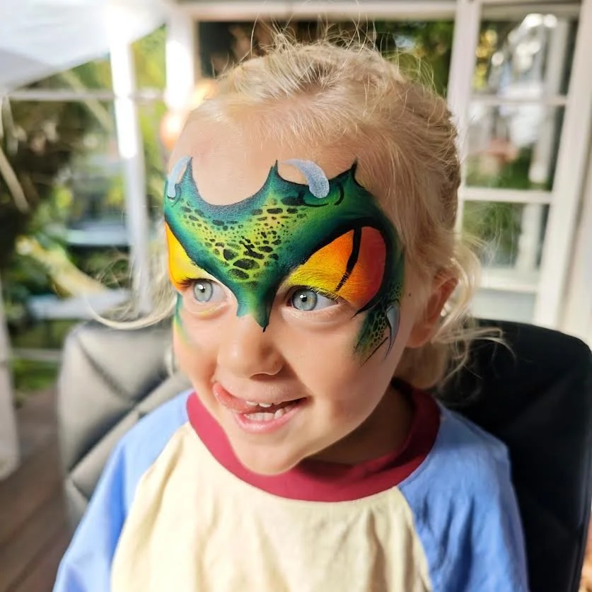 Young girl with face paint resembling a colorful dragon or dinosaur, smiling and looking to the side in a sunlit room.