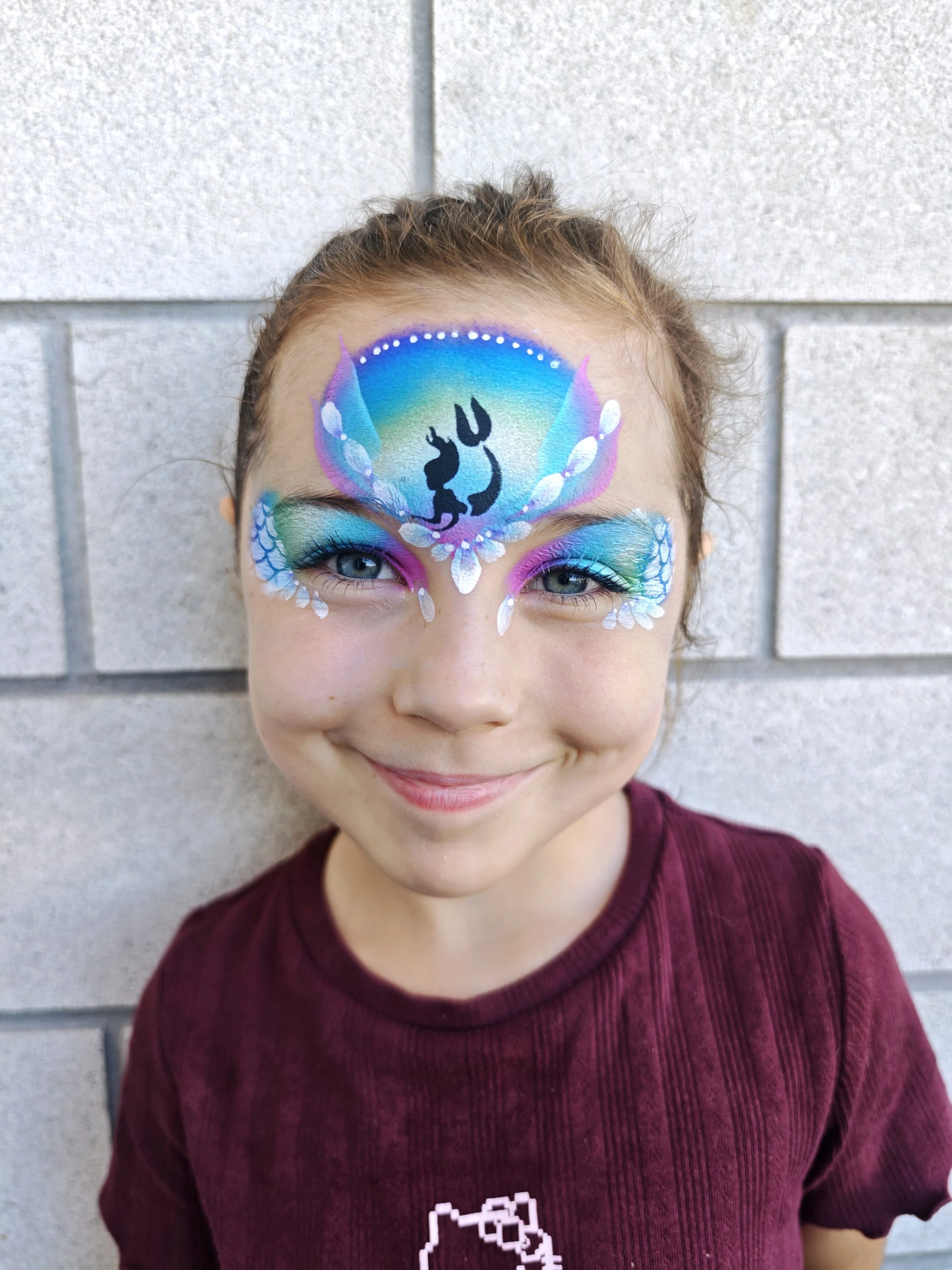 Young girl with an elaborate, colorful butterfly face paint design, featuring blue, purple, green, and white hues, with black silhouette of two cats in the center, standing in front of a gray brick wall.