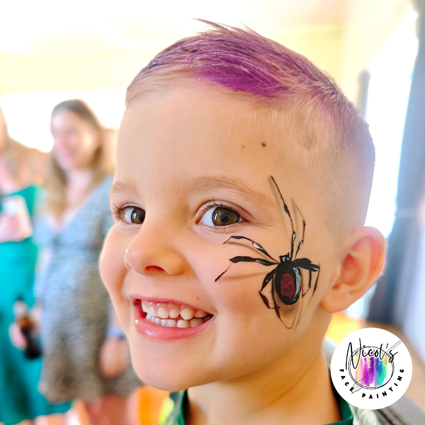A young boy with face paint of a black and red spider on his cheek, smiling at the camera, with other children in the blurred background.