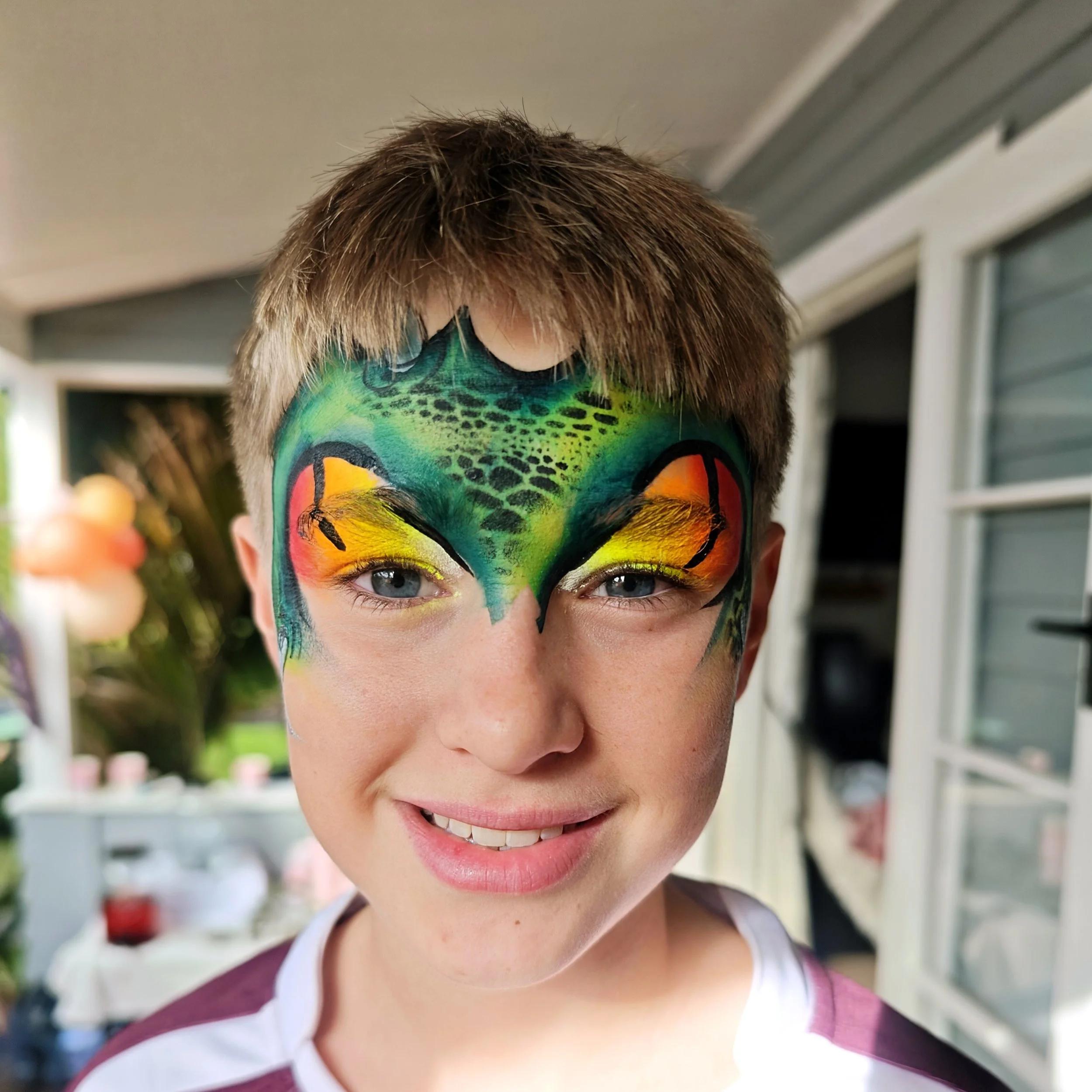 Close-up of a person's face with colorful face paint resembling a butterfly, blue eyes, short brown hair, and a smiling expression, outdoors near a window.
