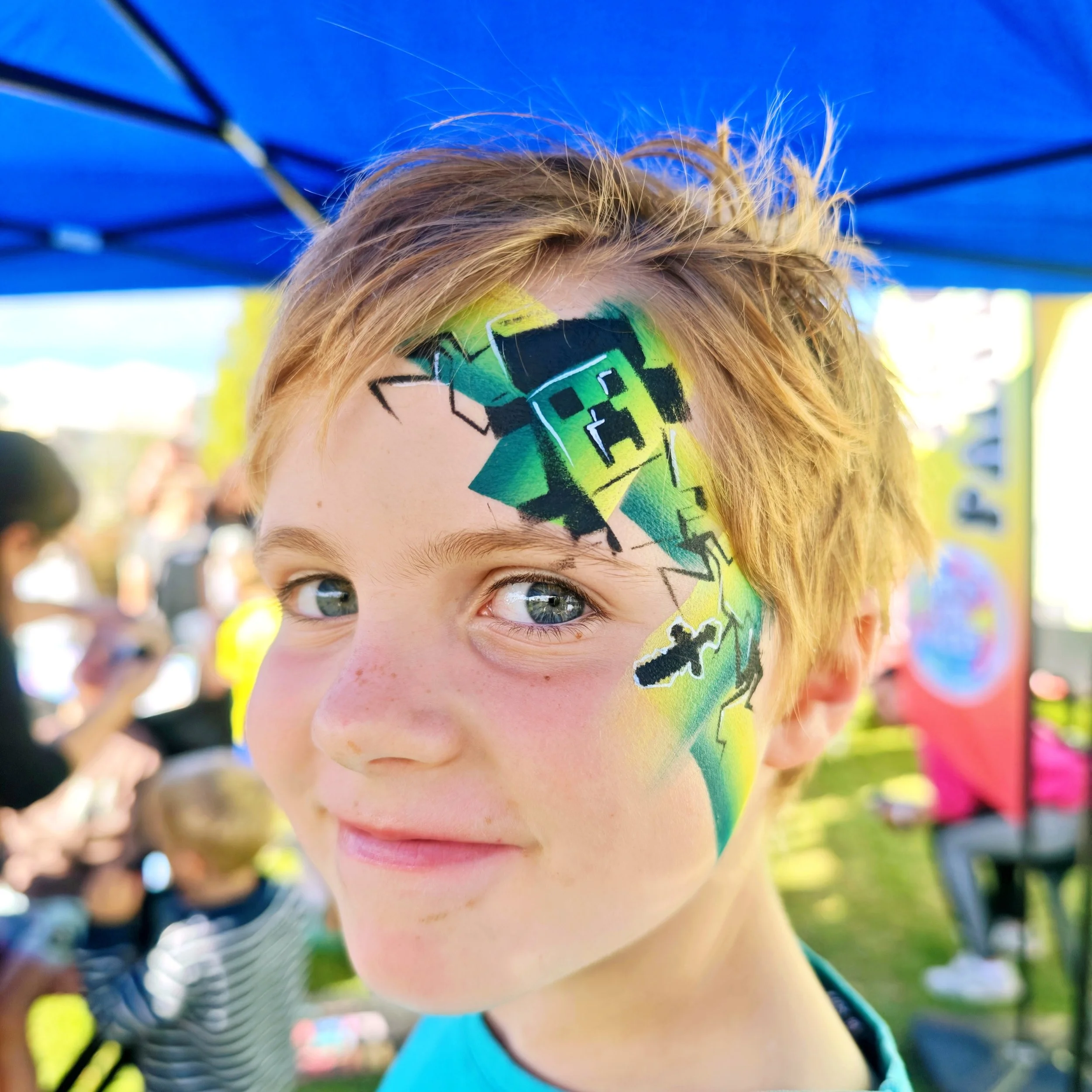 Close-up of a person's face with colorful geometric face paint, showing their blue eye and part of their nose, blonde hair, and a blue shirt, outdoors under a blue umbrella.