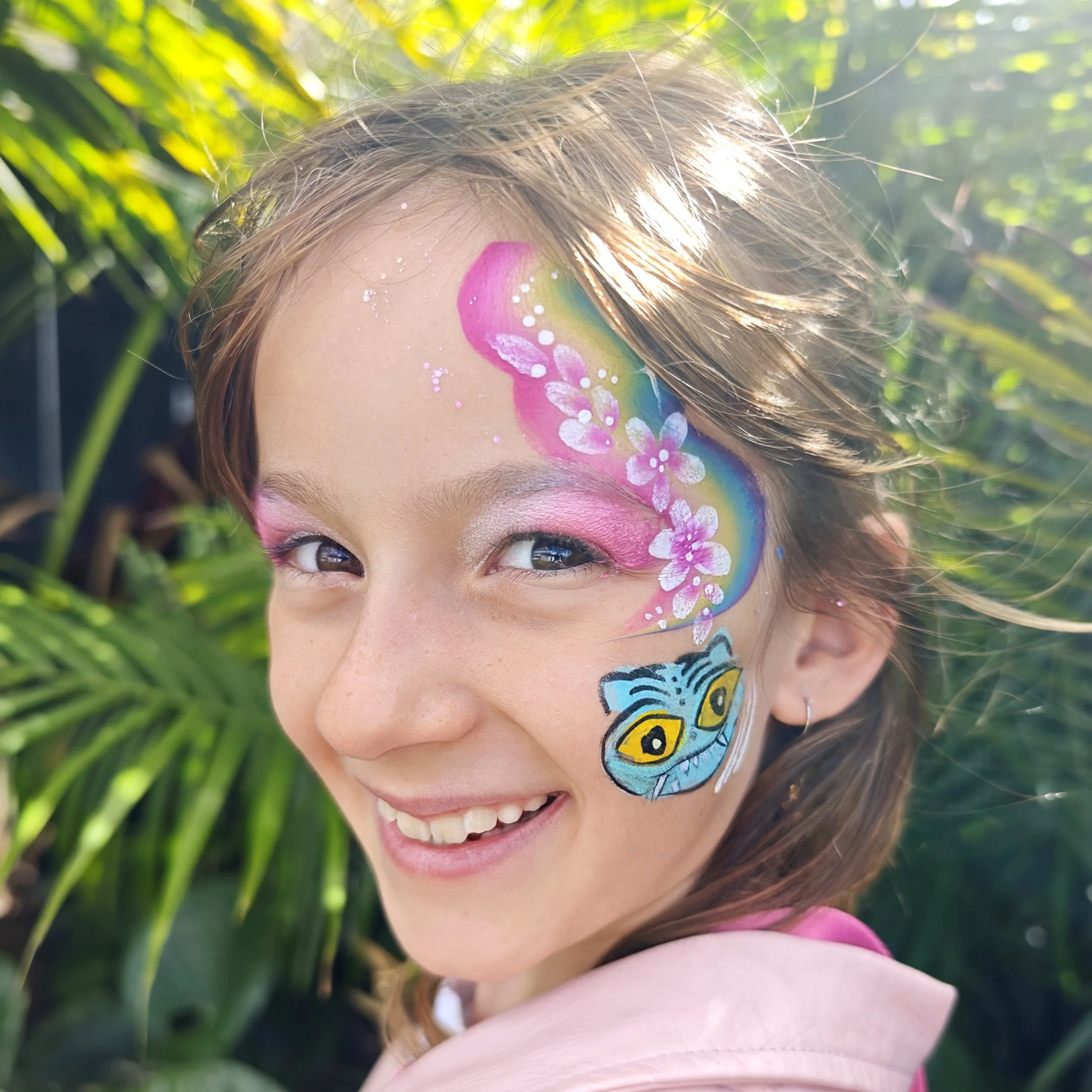 A young girl with face paint featuring a butterfly and floral designs, smiling outdoors with green foliage in the background.