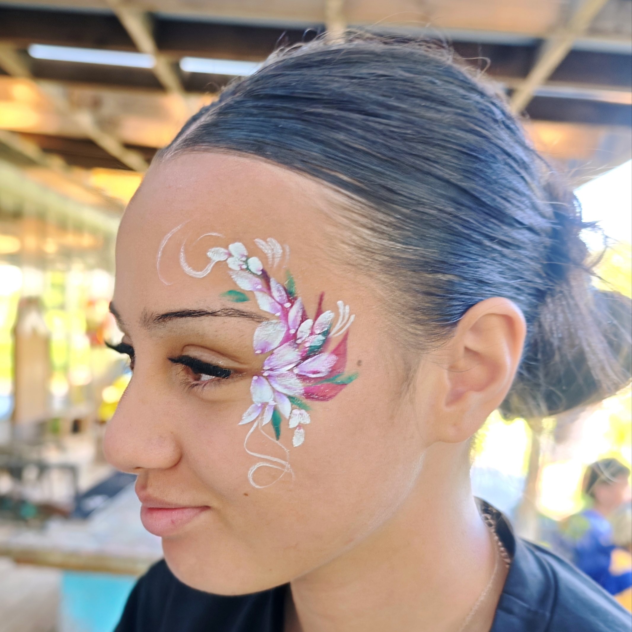 Close-up of a girl with painted flowers on her face and a braid hairstyle, wearing a dark shirt, outdoors with blurred background.