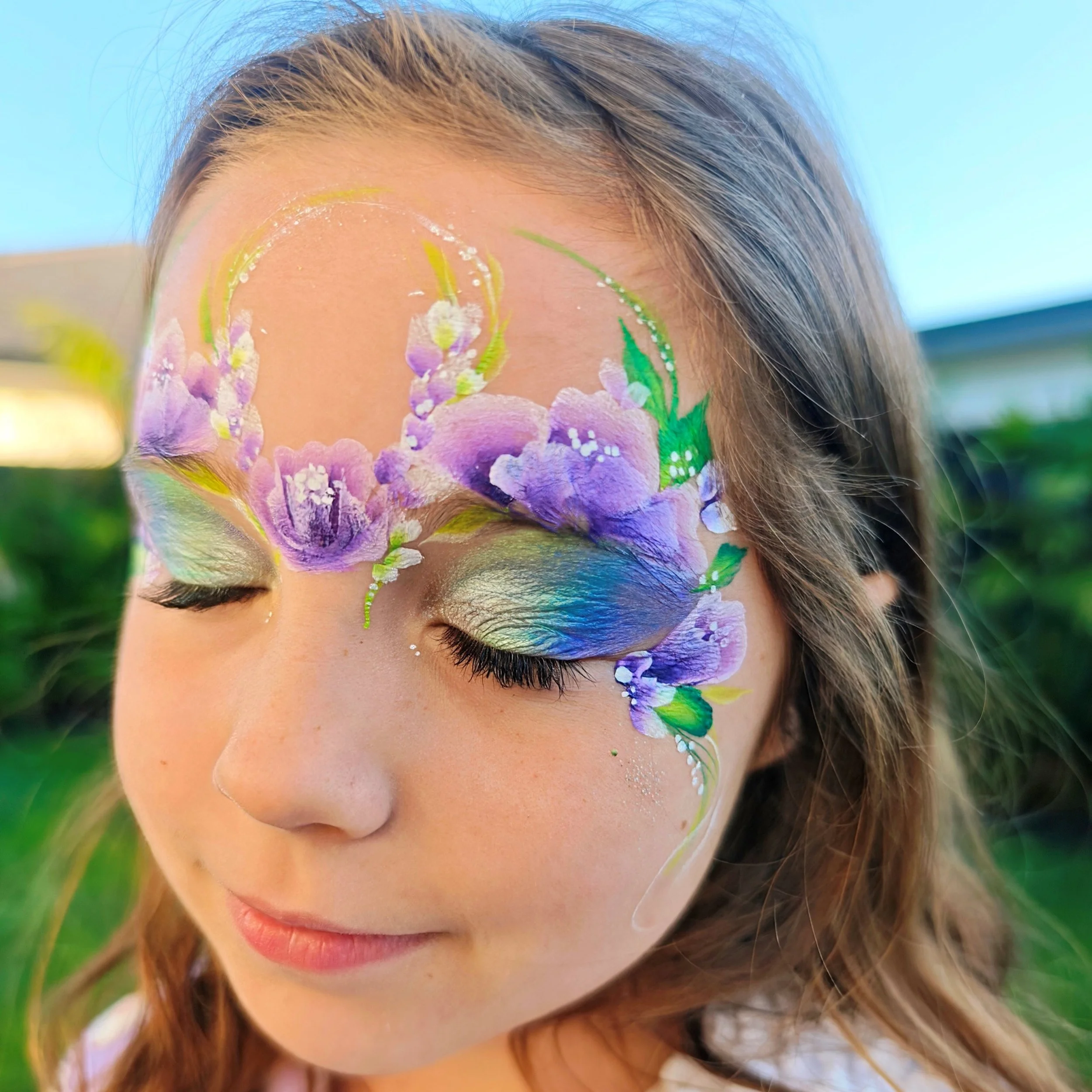 A person with painted face featuring purple, pink, and green floral designs around the eyes and forehead, with long brown hair and a closed-mouth expression.