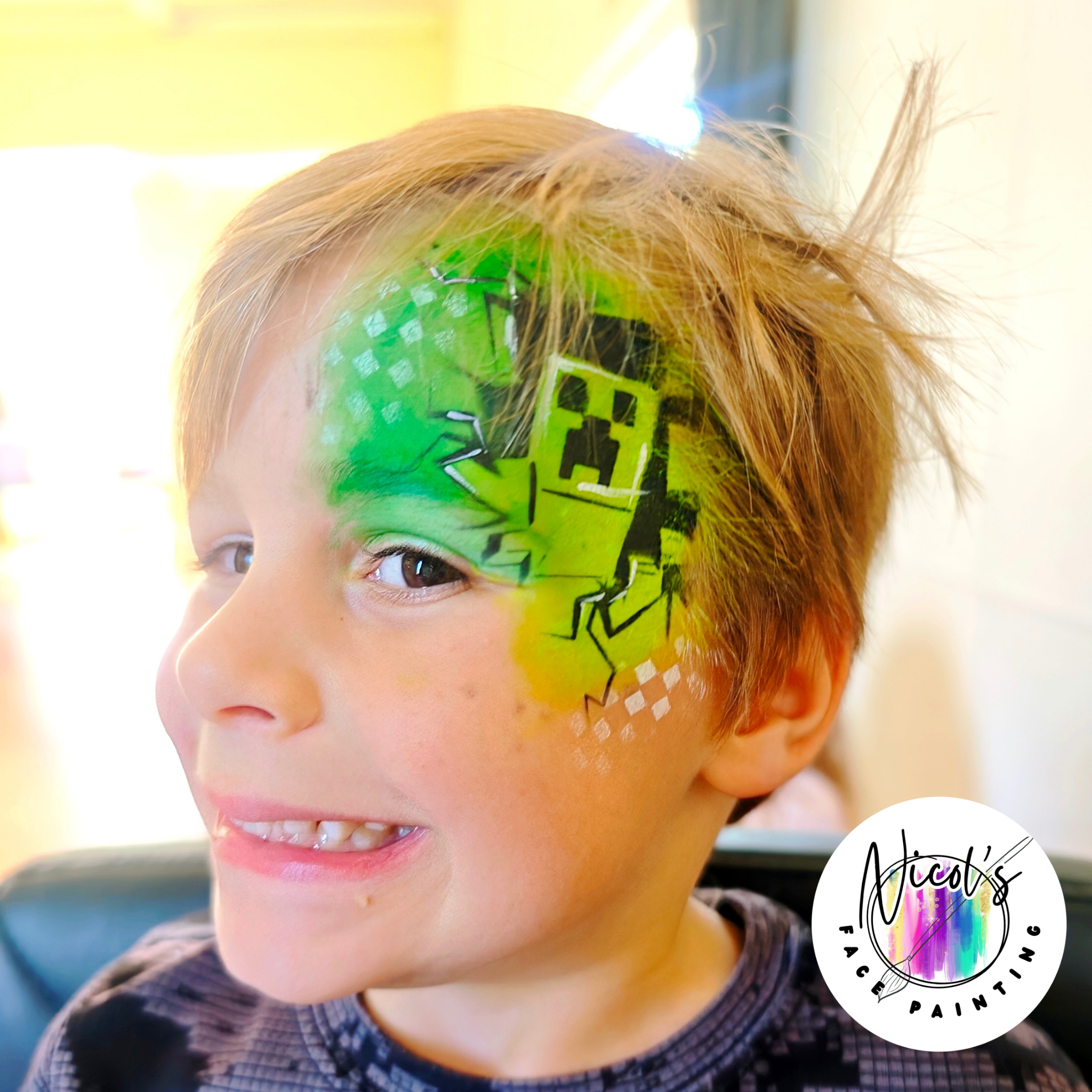 Close-up of a child's face with colorful face paint, including green, yellow, and black designs around the eyes and forehead. The child's brown hair is partially visible, and they are smiling.
