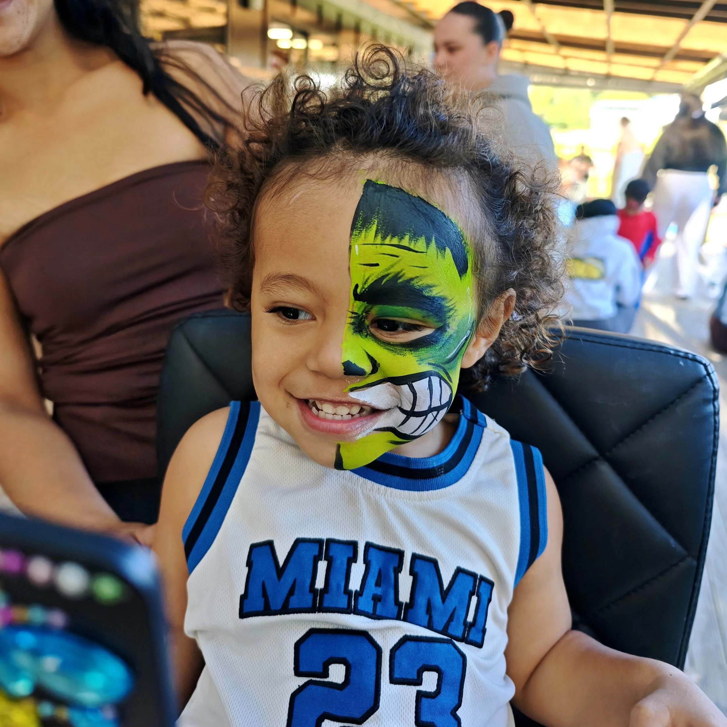 A young boy with face paint of a neon green and black skull is smiling. The background shows several people in a busy outdoor setting, possibly at a fair or festival.