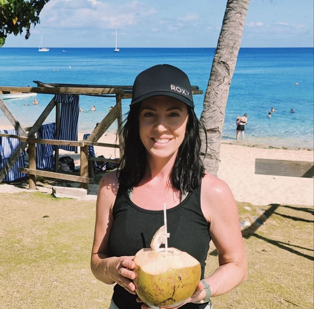 A woman with black hair wearing a black cap and tank top, smiling and holding a fresh coconut with a straw, on a beach with palm trees, blue ocean, and sailboats in the background.