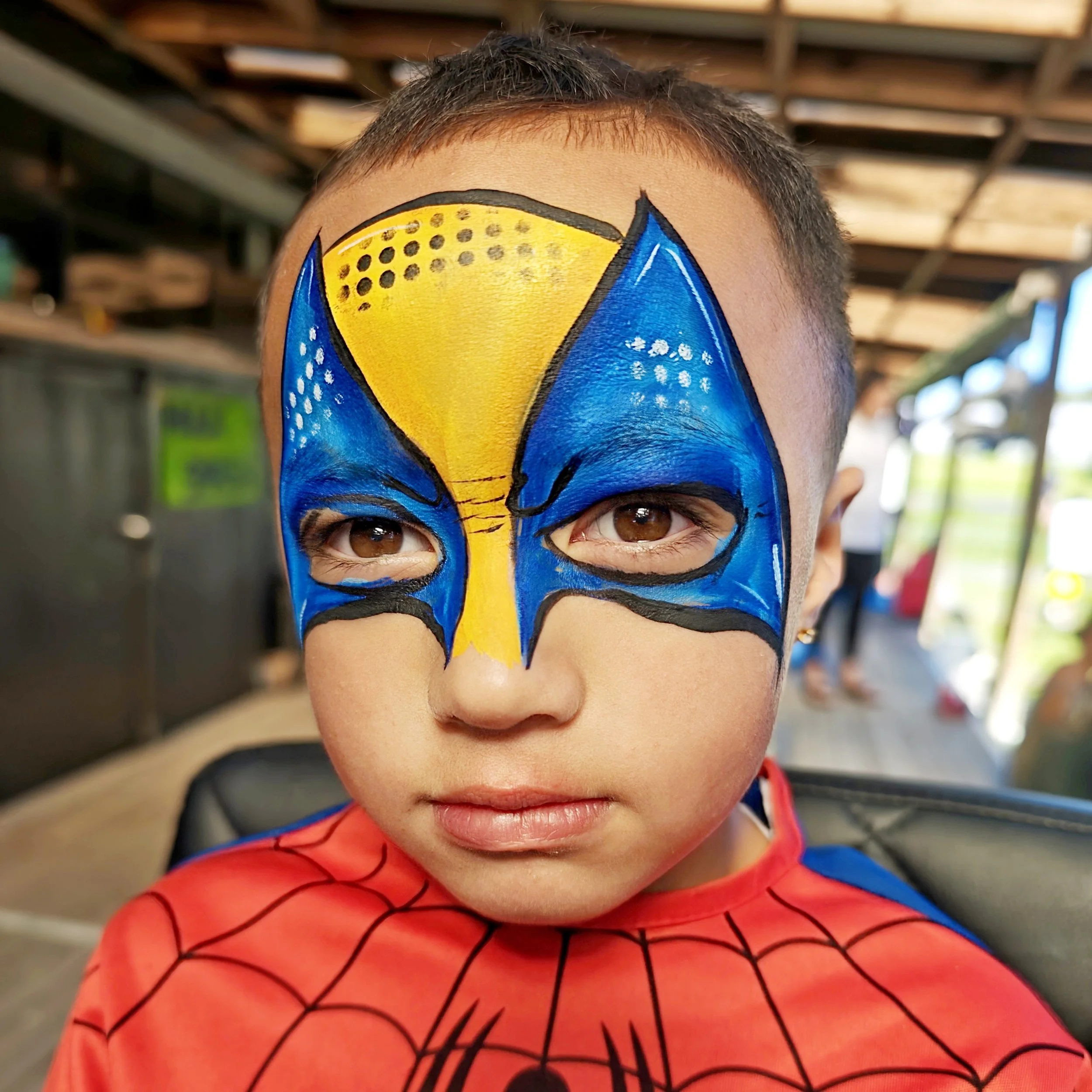 Close-up of a young boy with face painted as Spider-Man, wearing a red and blue Spider-Man costume, indoors.