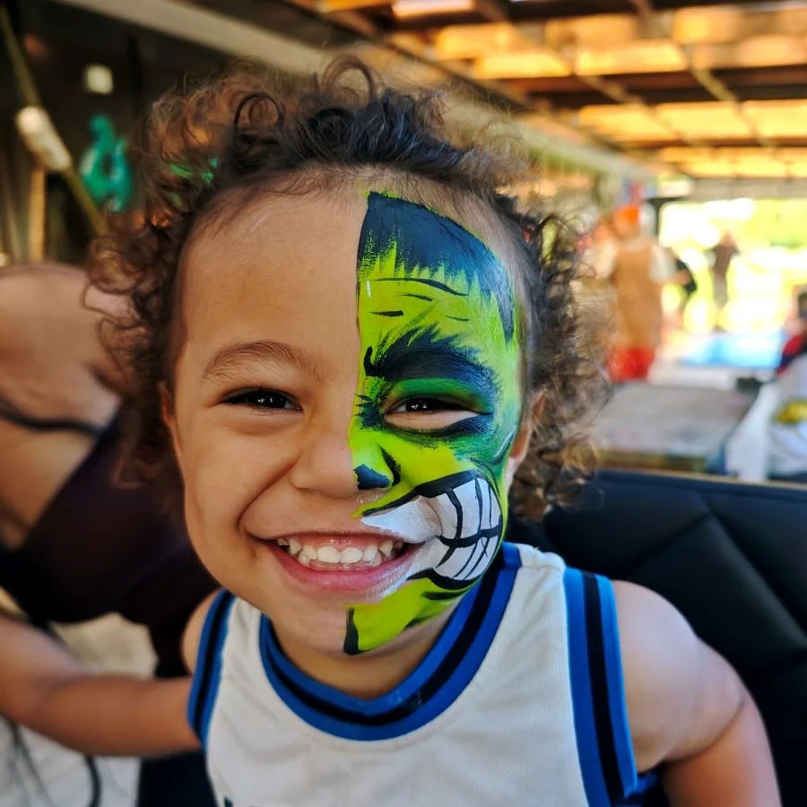 Young boy with face paint of a green and blue tiger, smiling cheerfully.