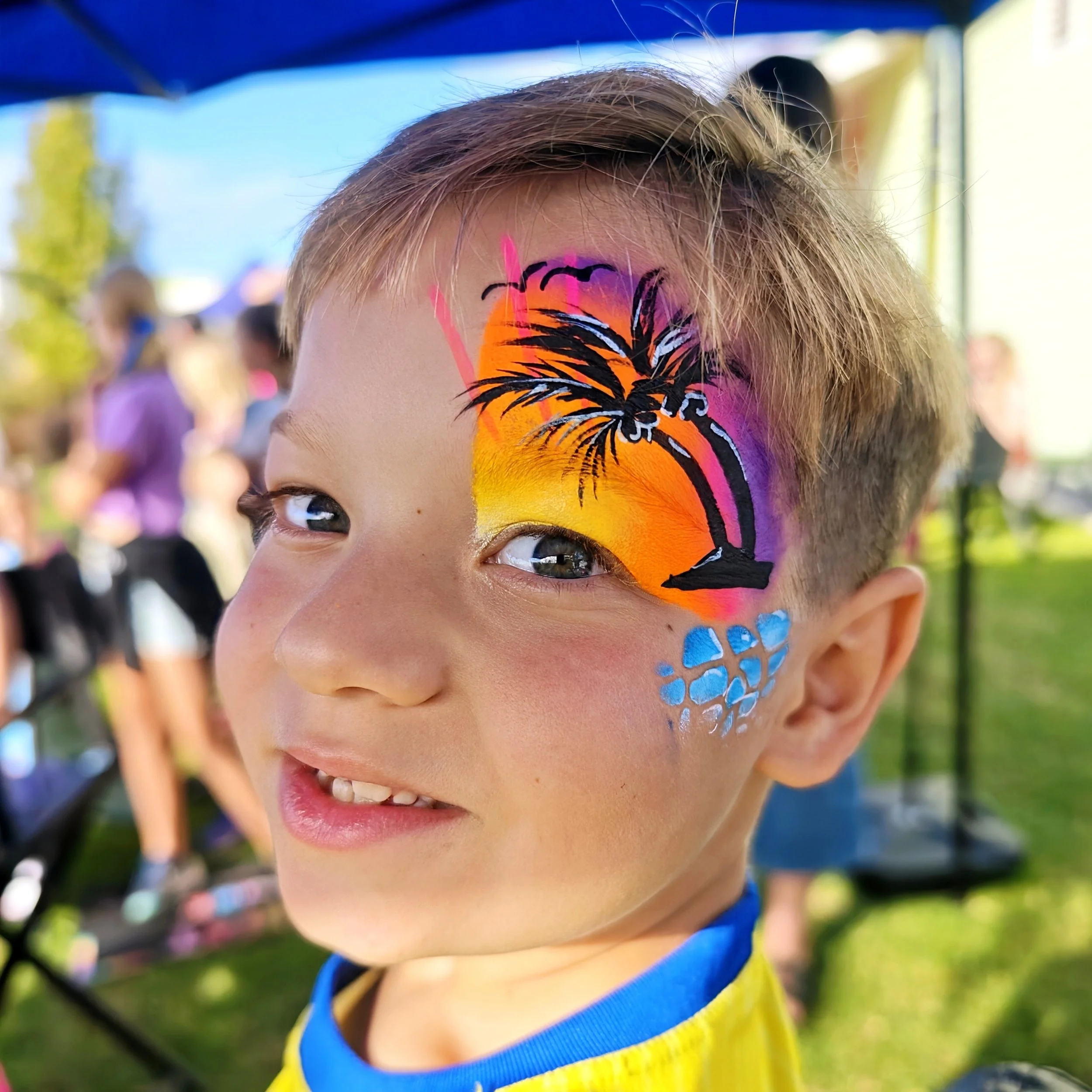 Child with face painted with vibrant sunset colors and black palm tree silhouette, outdoors, smiling, wearing a blue and yellow outfit.