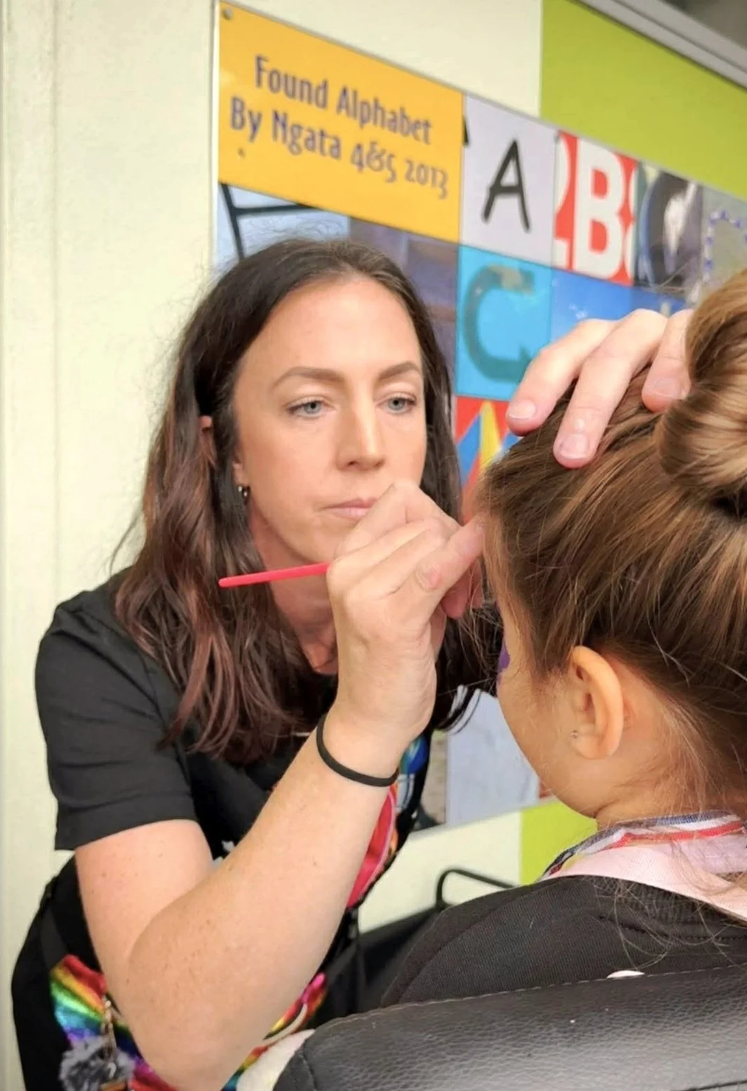 A woman with dark brown hair paintings or drawing on the face of a young girl with brown hair tied in a bun. The woman is holding a small paintbrush and has her hand gently placed on the girl's head. In the background, there's a colorful educational display with letters and a sign that reads "Found Alphabet By Ngata 4 Dec 2013".