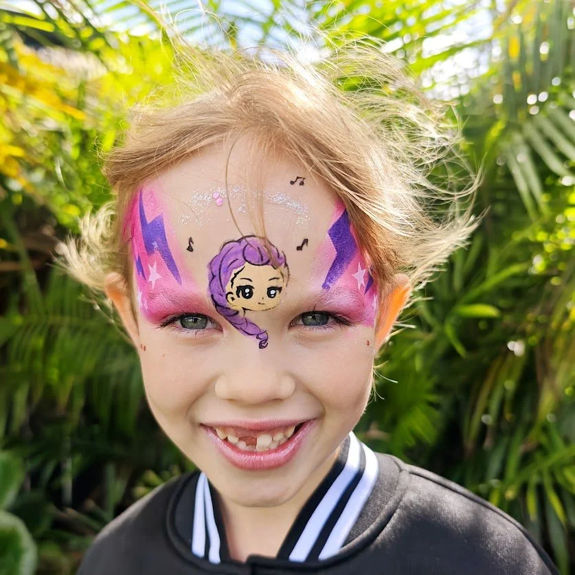 Young girl with face paint featuring a cartoon character with purple hair, surrounded by pink, purple, and blue designs, outdoors with green foliage in the background.