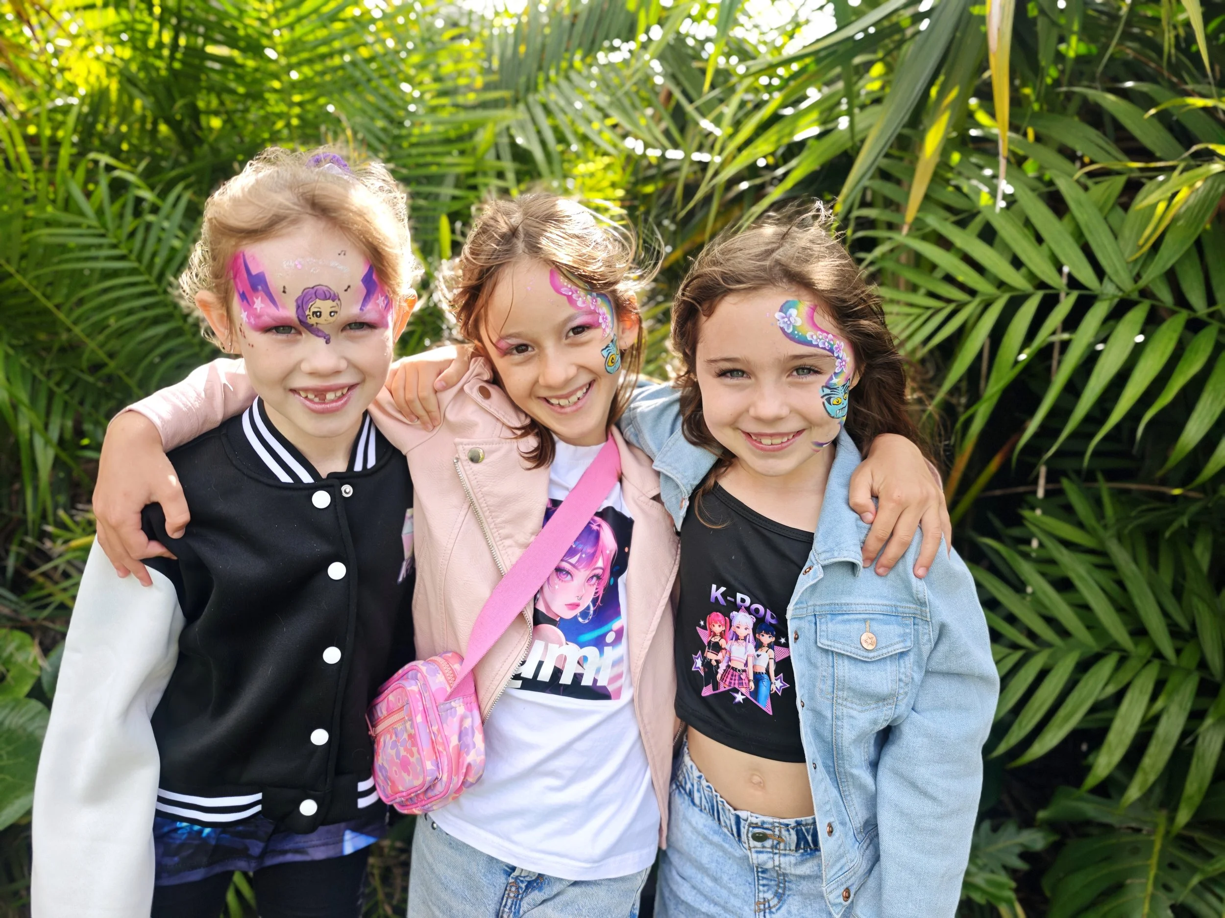 Three young girls with face paint smiling and hugging each other outdoors with green tropical plants in the background.