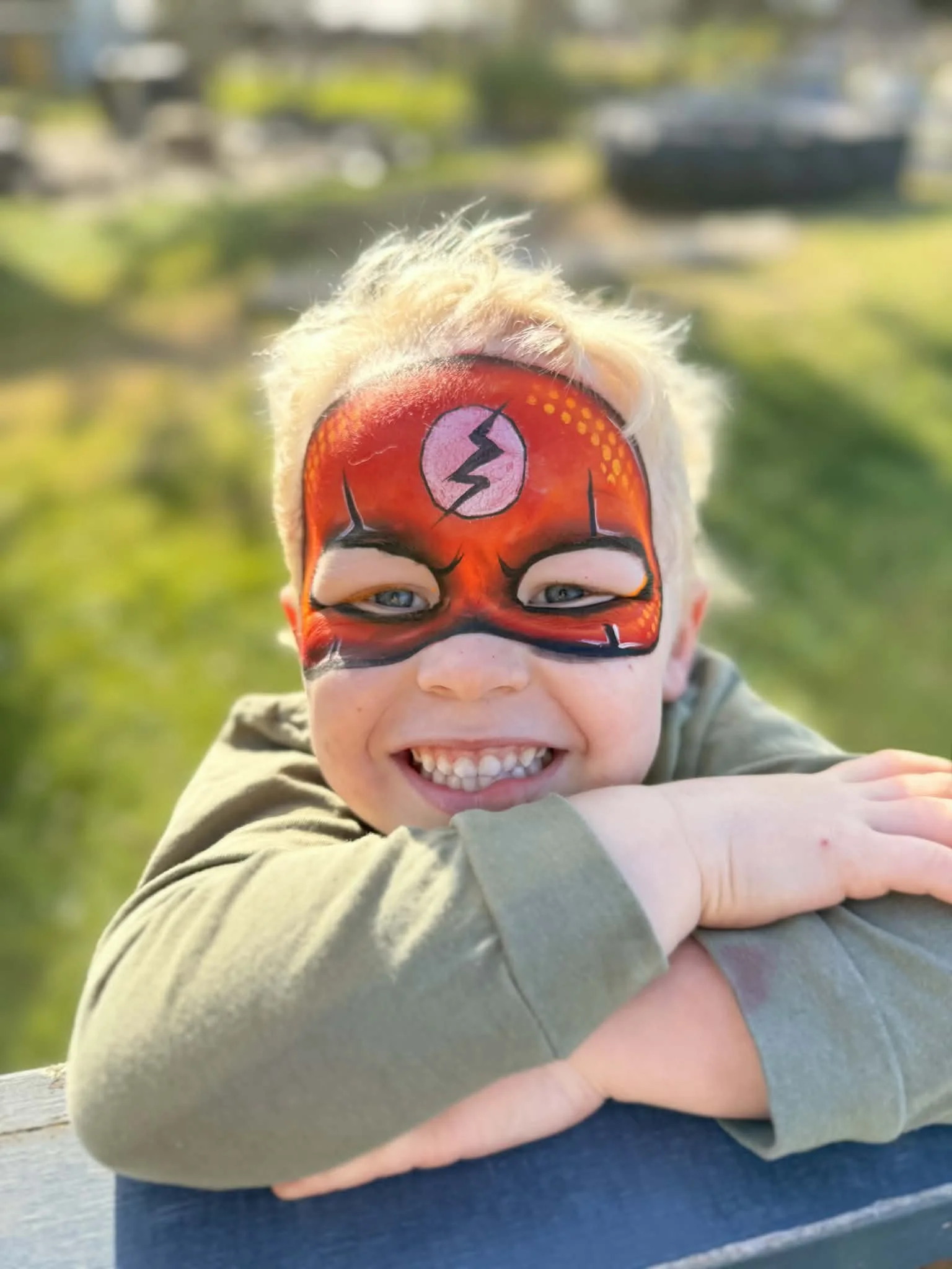 Child with face painted as a superhero, showing a lightning bolt symbol and a pink circle on forehead, hugging an adult outdoors.