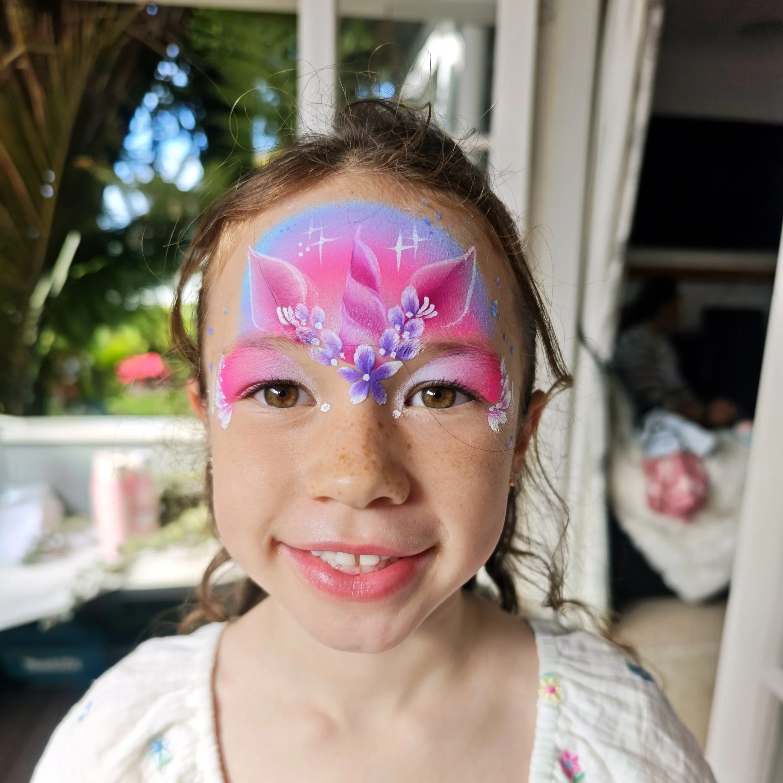Close-up of a young girl with face painted with a colorful floral design, smiling in an outdoor setting.