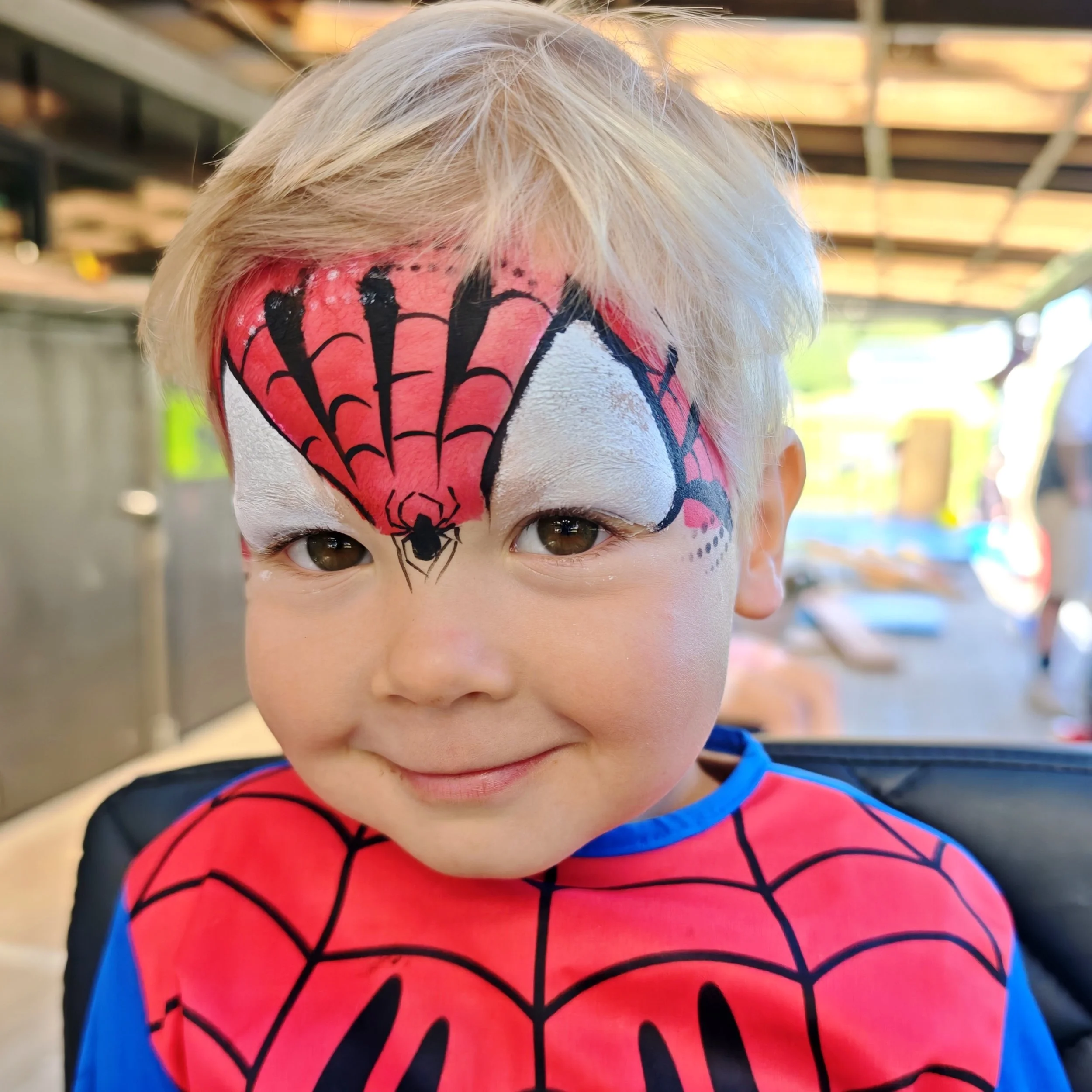 Close-up of a child's face painted as Spider-Man, with blonde hair and brown eyes, wearing a Spider-Man costume with a red and blue web pattern, outdoors under a wooden structure with sunlight in the background.