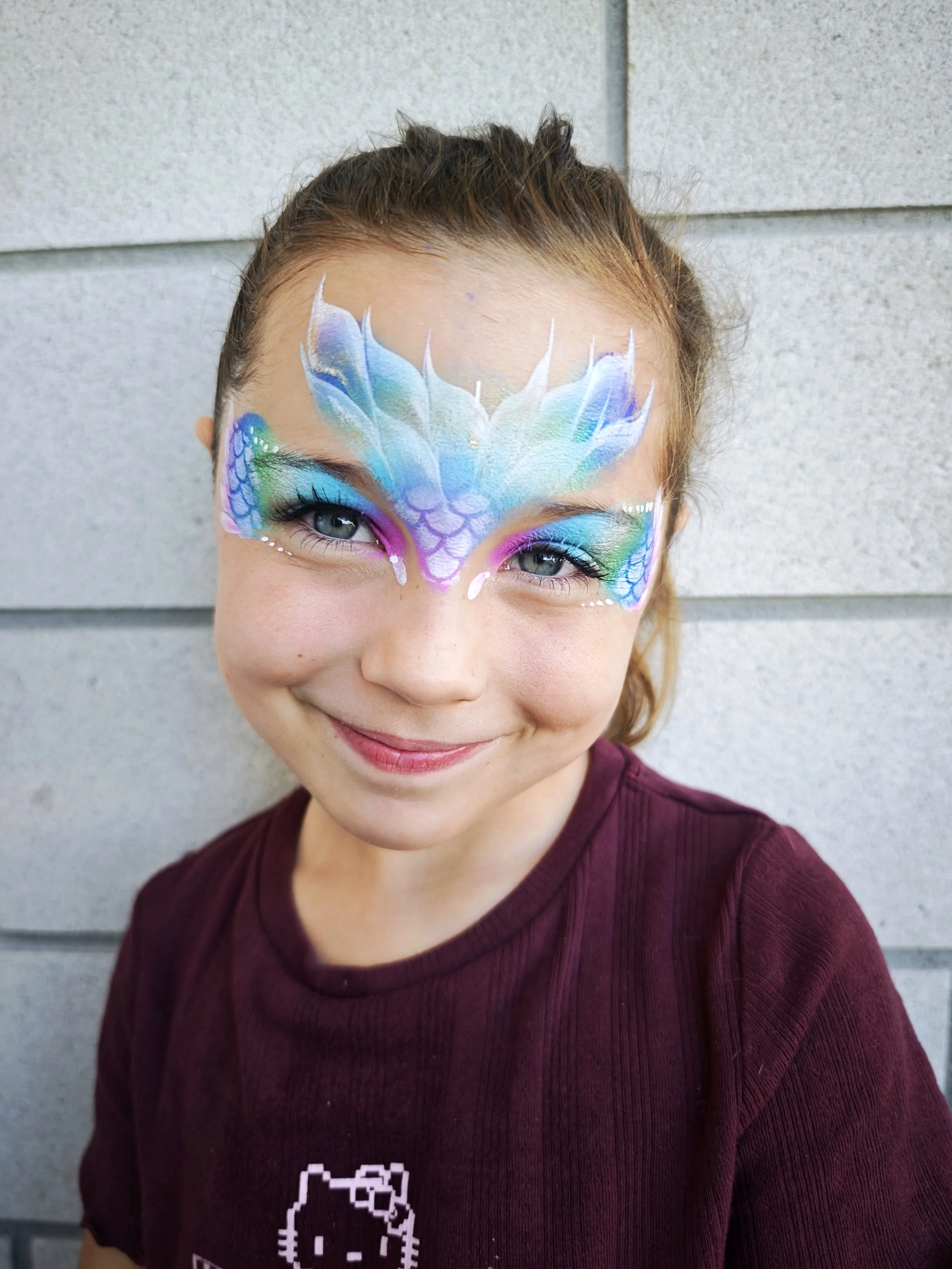 A person with face paint resembling a colorful, detailed dragon on their face, smiling, wearing a maroon sweater with a pixel art character on it, standing in front of a gray wall.