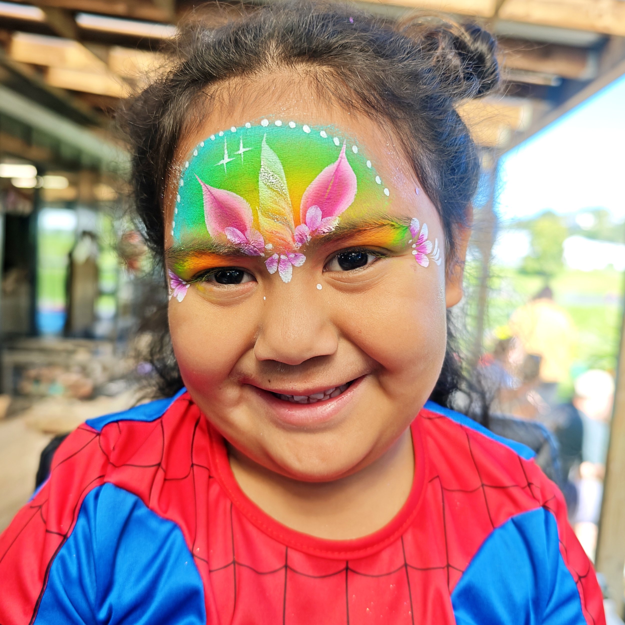 A young girl with curly hair smiling, wearing a colorful face painting of pink lotus flowers and green leaves on her forehead, dressed as Spider-Man with a red and blue costume.