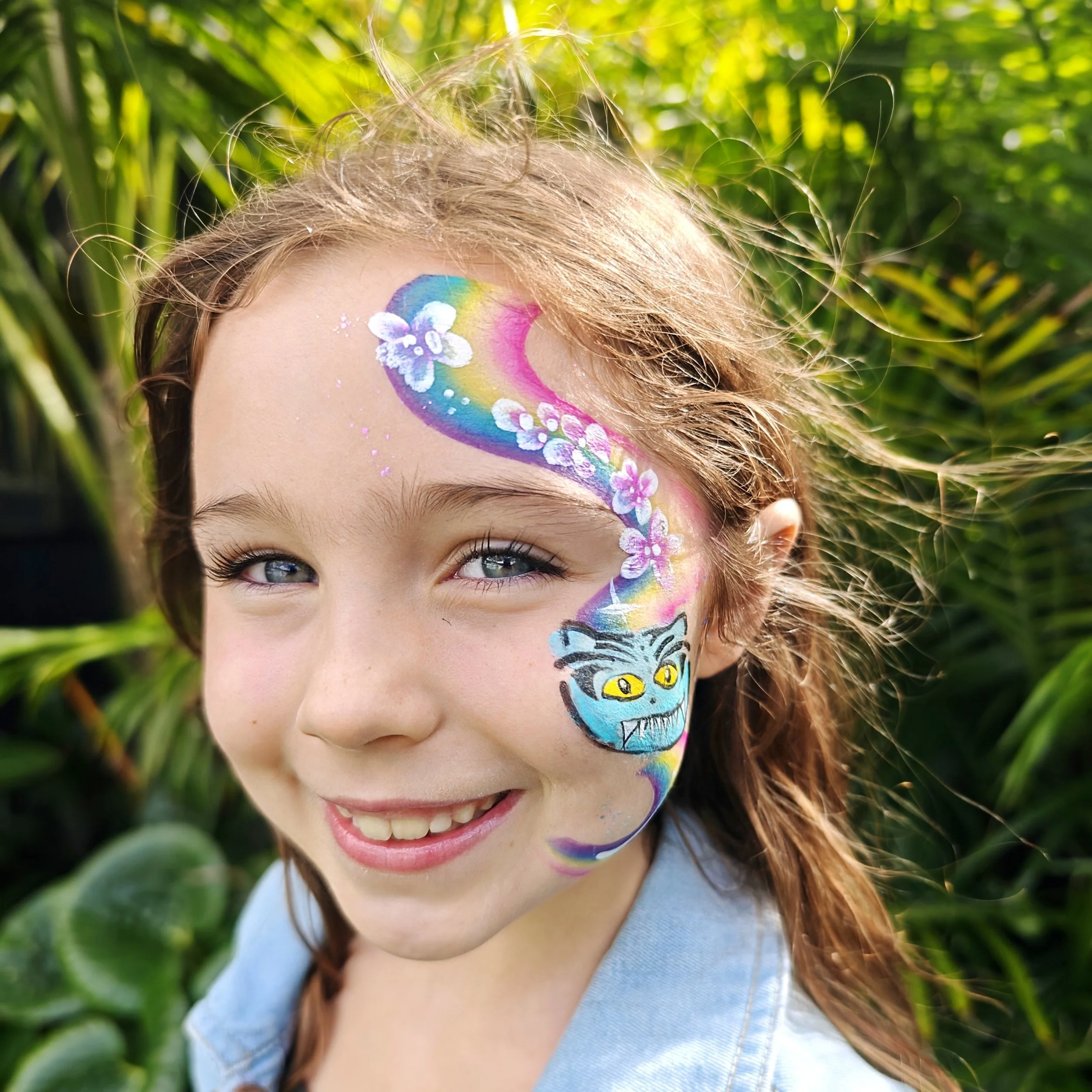 Close-up of a girl with face paint depicting a rainbow, flowers, and a blue skull with yellow eyes, outdoors with greenery in the background.