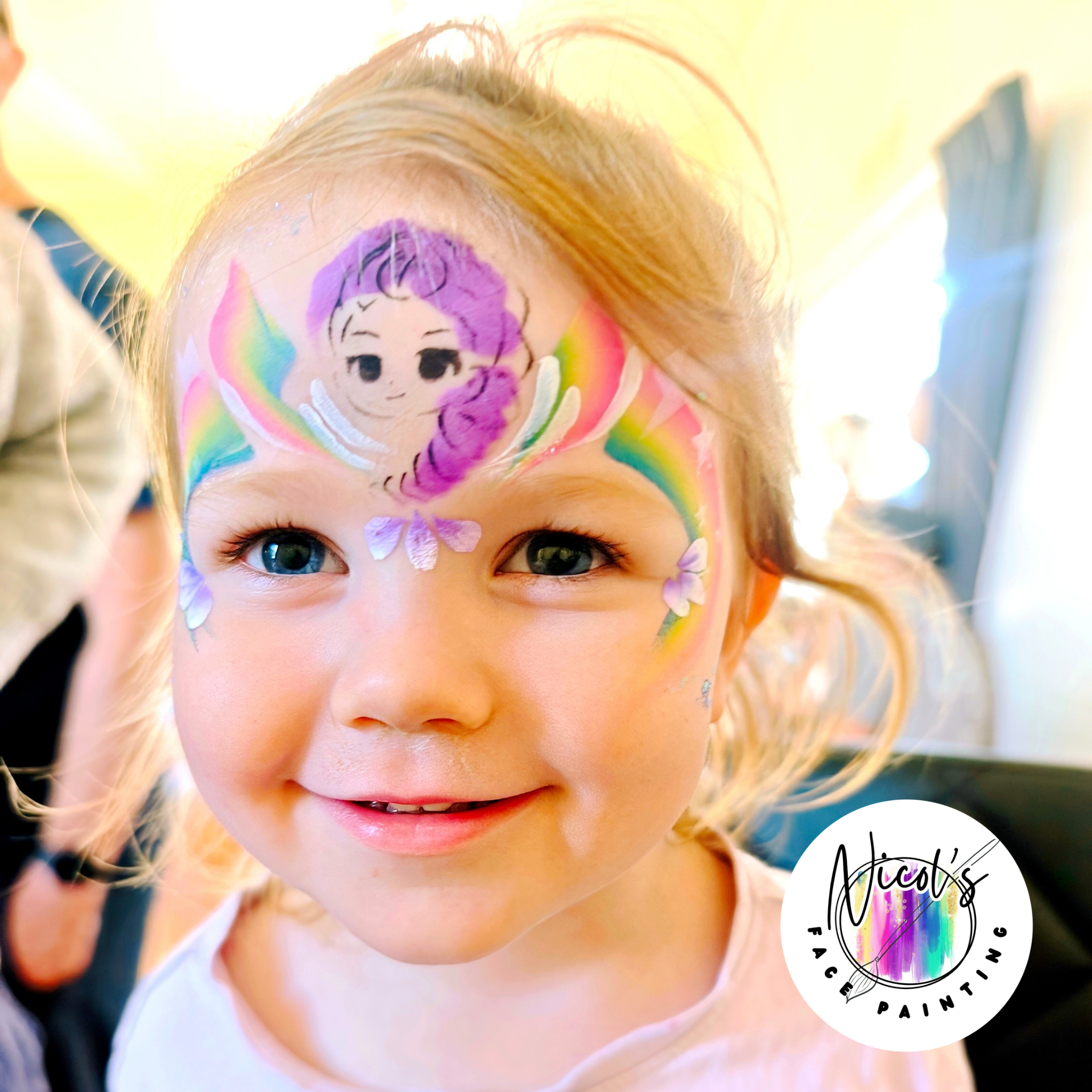 Close-up of a child's face with colorful face paint design featuring a purple and rainbow unicorn with big eyes and a rainbow mane, with light hair and bright eyes.