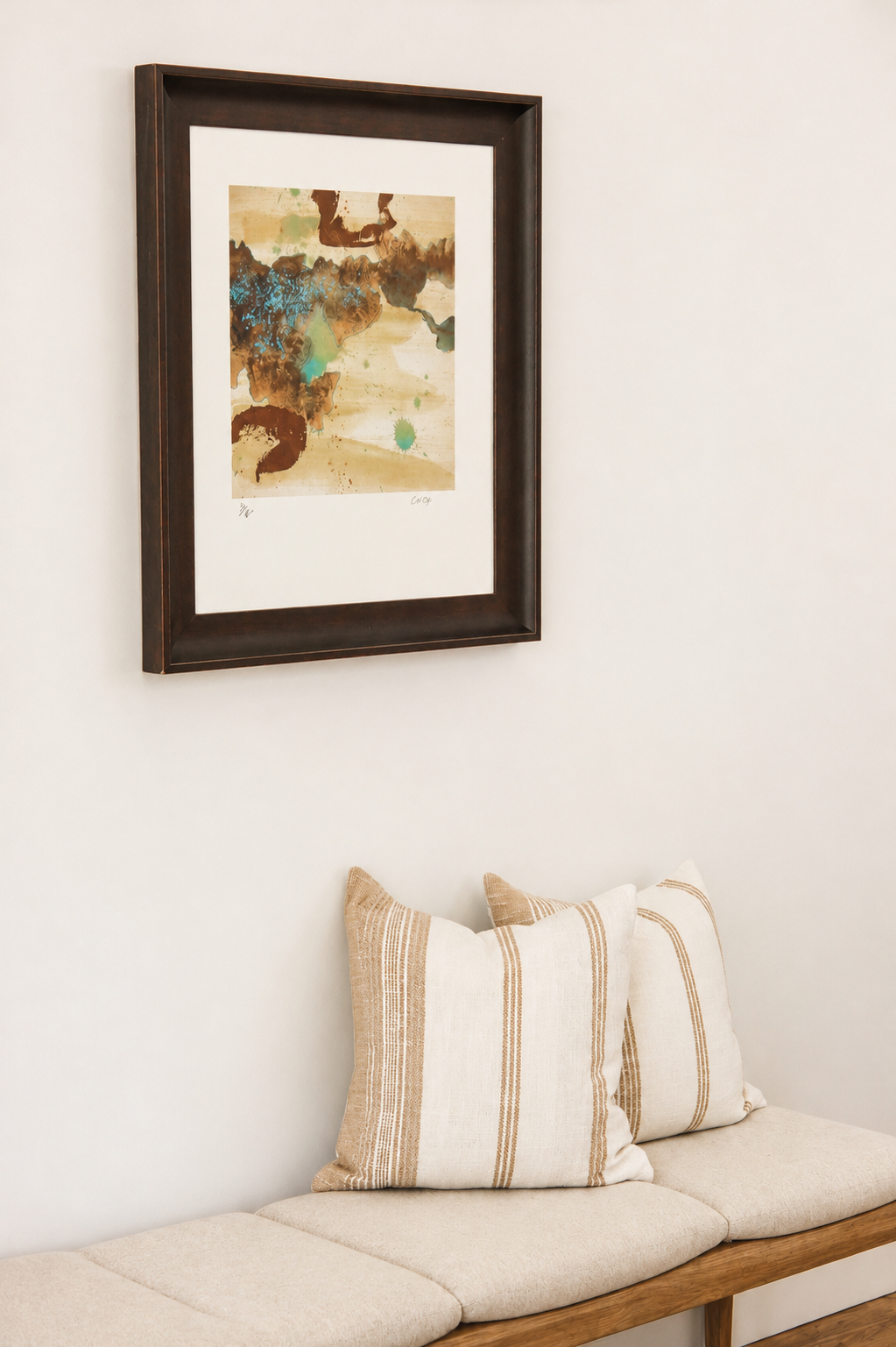 A framed abstract watercolor painting hanging on a white wall above a beige bench with striped pillows.