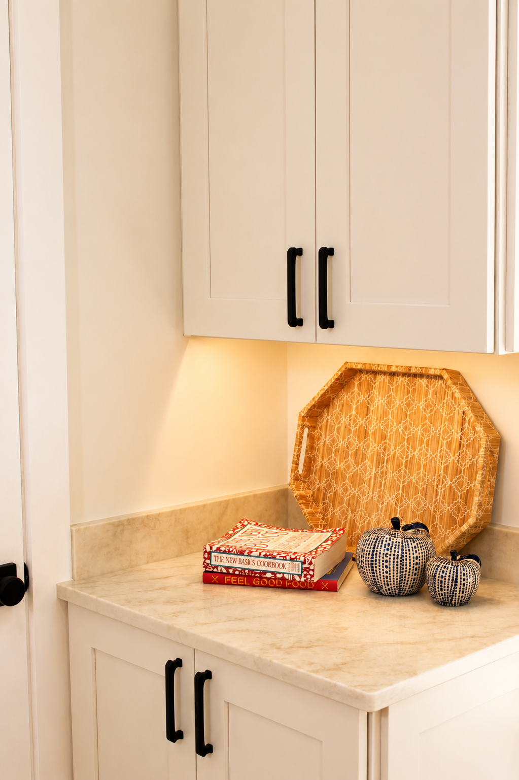 Kitchen countertop with two patterned ceramic jars, a tray, and two recipe books, against a white wall with upper and lower cabinets with black handles.