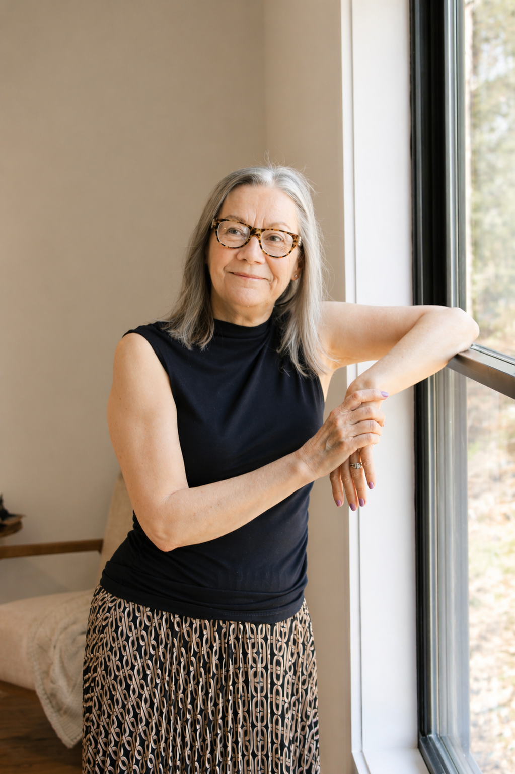 An older woman with glasses and shoulder-length gray hair standing by a window, wearing a sleeveless black top and patterned pants, smiling gently.