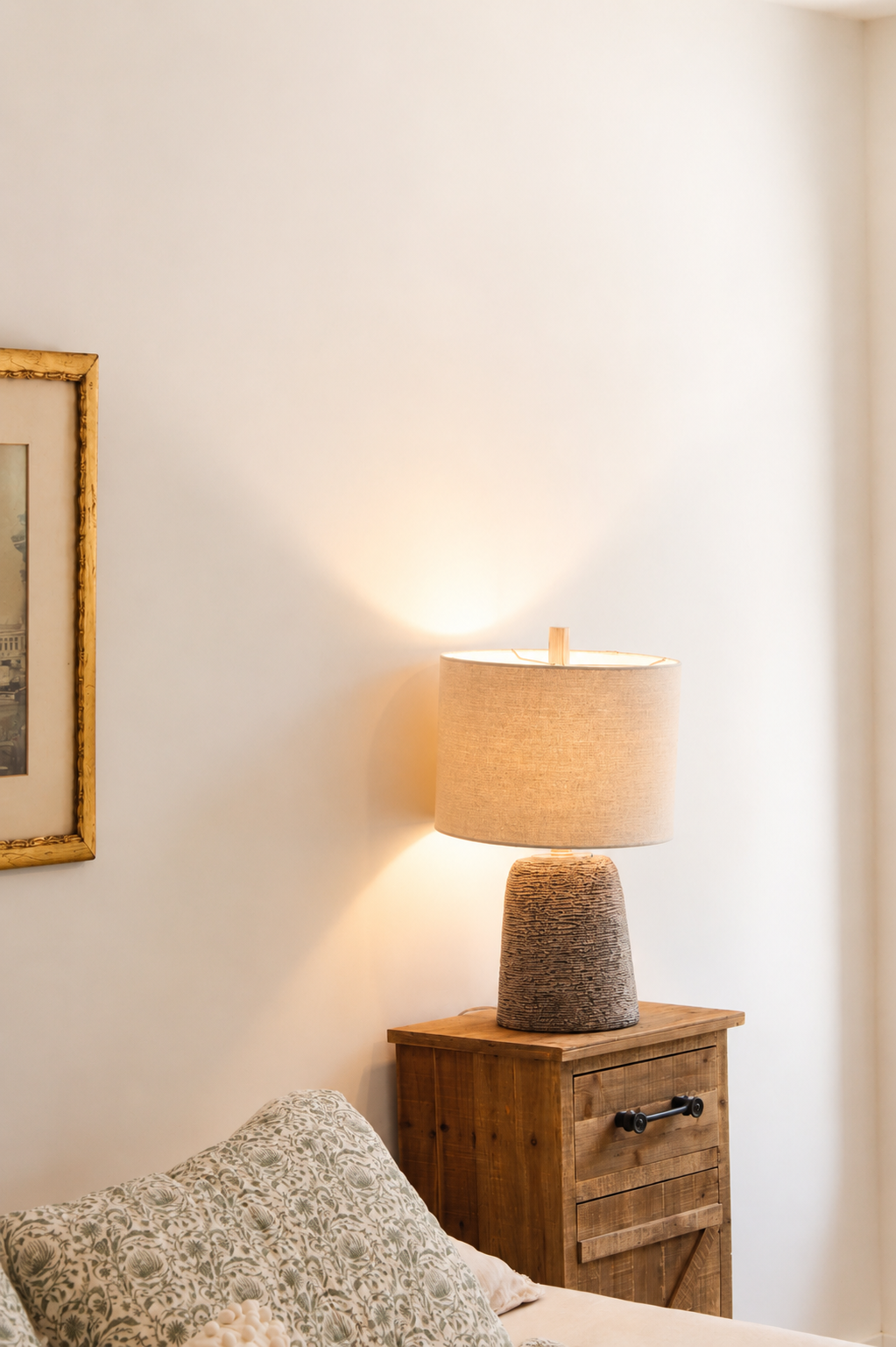 A cozy bedroom corner with a wooden nightstand, a textured table lamp, a patterned pillow, part of a bed, wall art, and a plain white wall.