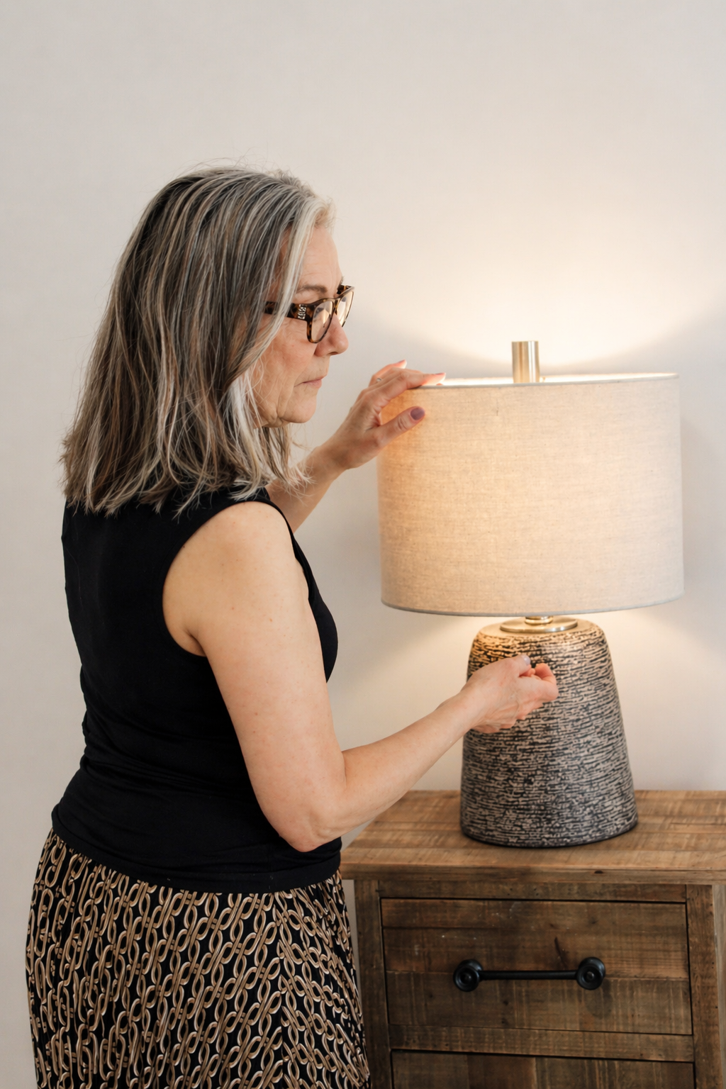 An elderly woman with gray hair and glasses adjusting a beige lampshade on a table lamp, standing next to a wooden cabinet.