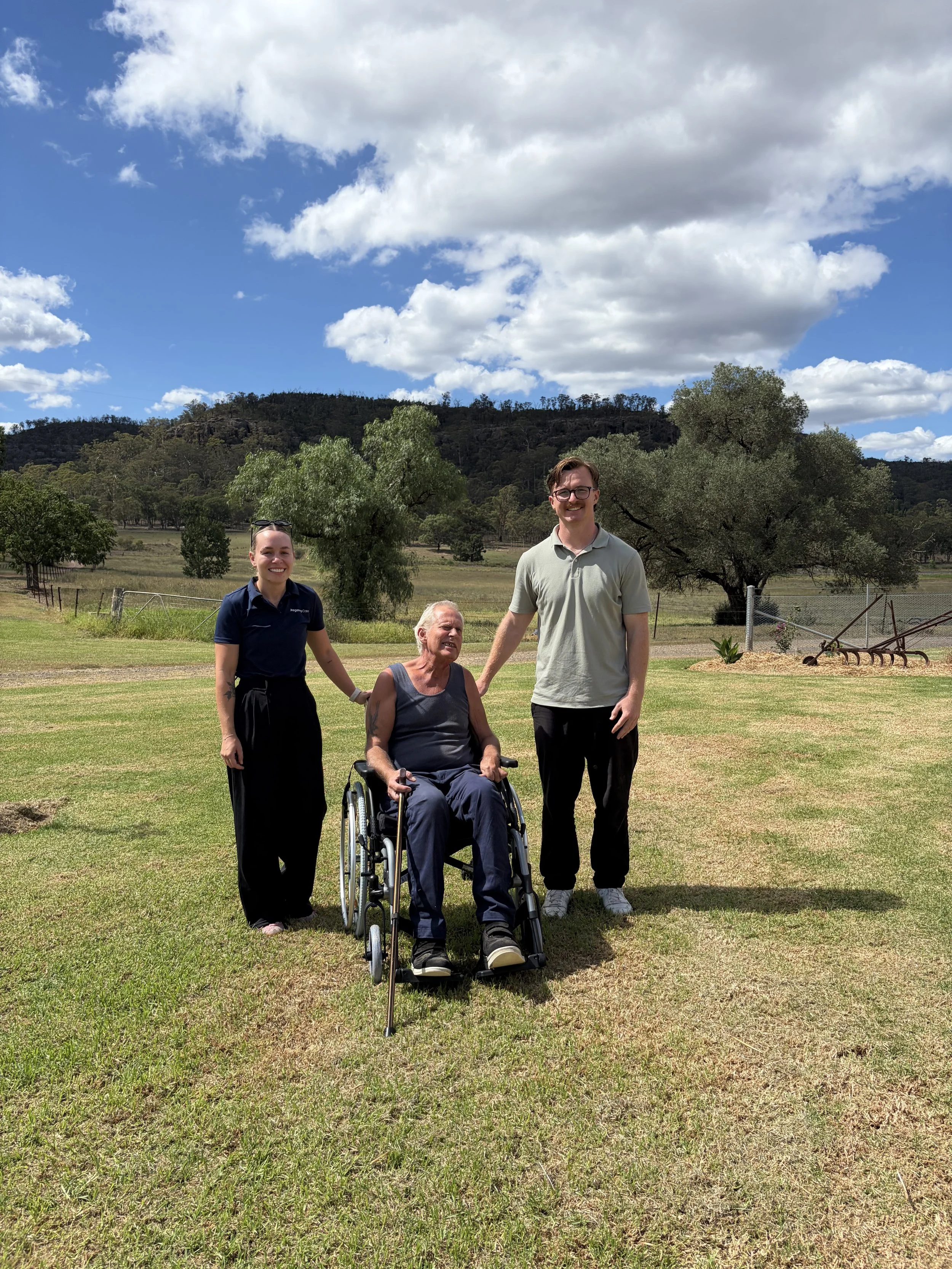 Four people standing on grass outdoors with trees and hills in the background, one person in a wheelchair, all smiling, under a partly cloudy sky.