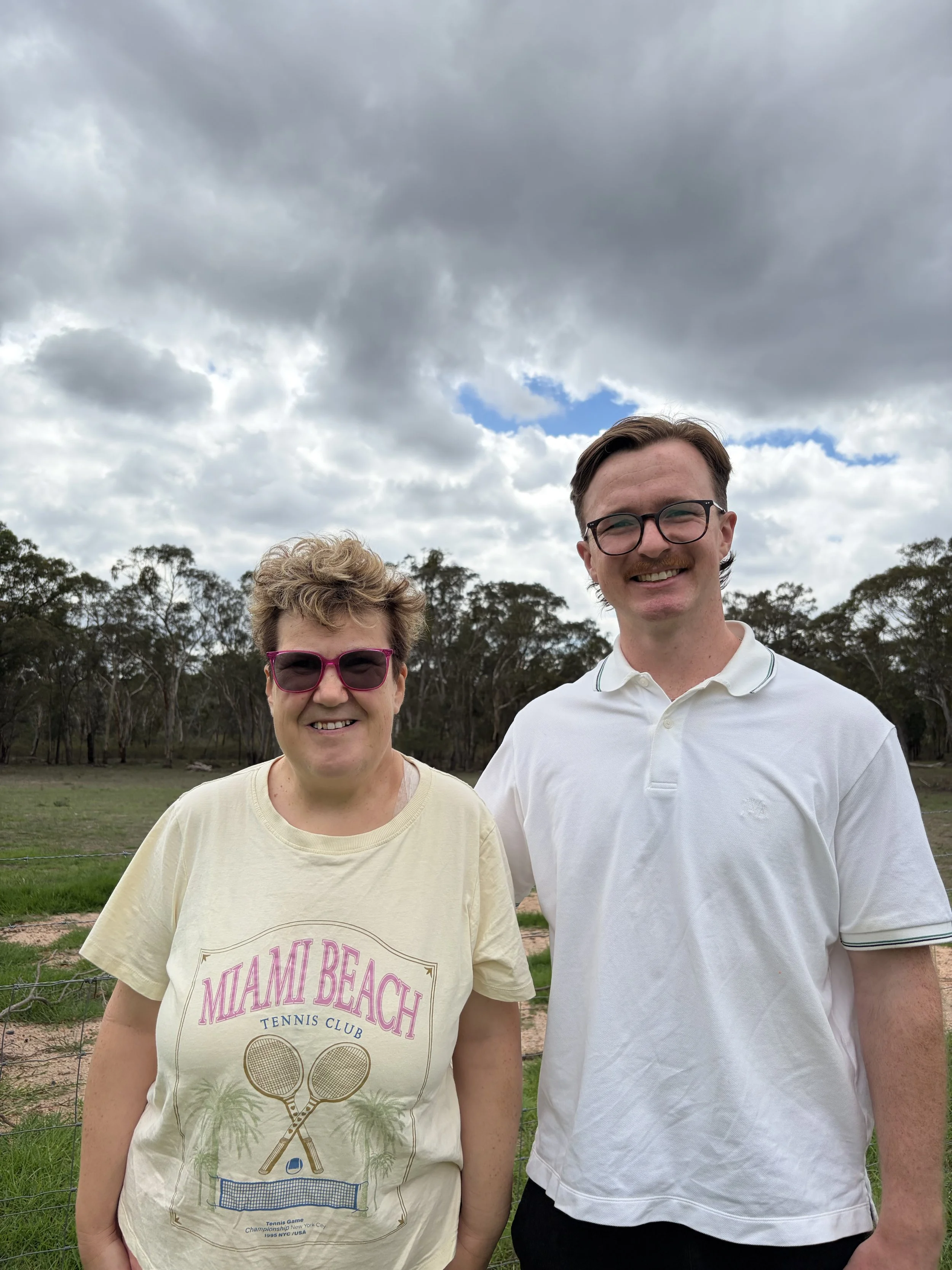 A woman and a man stand outdoors under a cloudy sky, smiling at the camera. The woman is wearing pink sunglasses and a yellow T-shirt with 'Miami Beach Tennis Club' written on it. The man has short brown hair, glasses, and is wearing a white polo shirt.