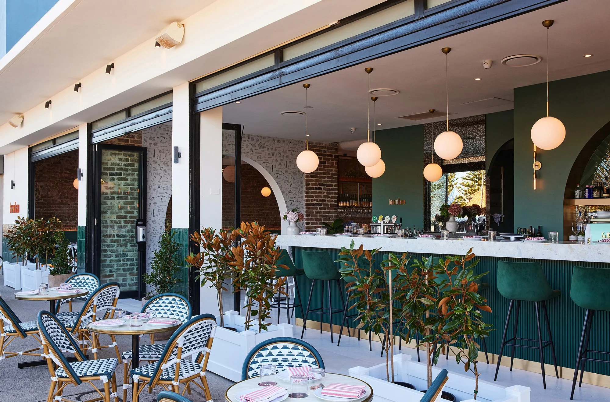 Inside view of a stylish restaurant or cafe with a bar counter, green chairs, potted plants, hanging round pendant lights, and a large window showing outdoor scenery.
