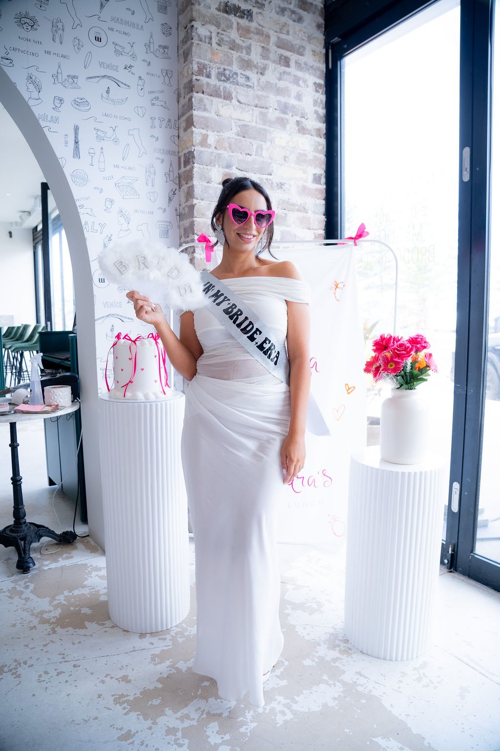 Woman wearing a white dress, pink heart-shaped sunglasses, and a sash that says 'Bride Tribe.' She is standing in front of a white floral backdrop with a bouquet of pink flowers on a white pedestal. She is holding a white fan.