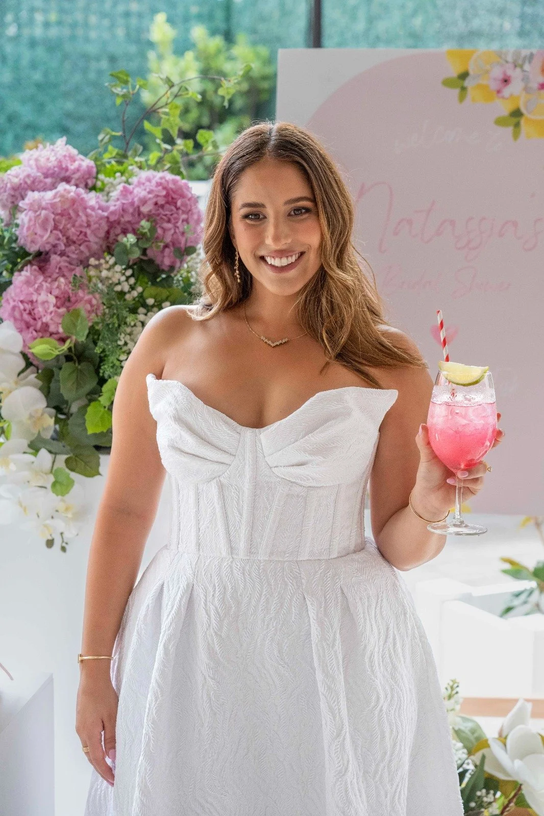 A woman in a white strapless dress holding a pink cocktail with a lemon wedge and a striped straw, standing in front of floral decorations and a pink sign.