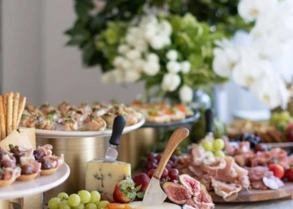 Buffet table with assorted cheeses, grapes, strawberries, figs, cold cuts, and appetizers, with a bouquet of white flowers in the background.