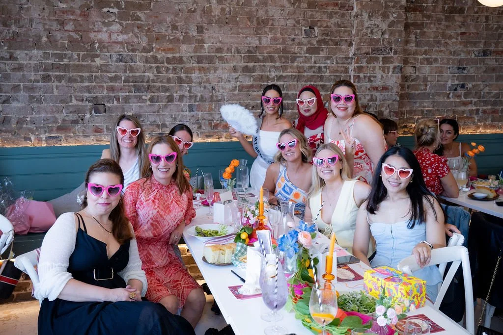 Group of women at a brunch or party wearing pink heart-shaped sunglasses, sitting and standing around a decorated table with food and drinks in a brick-walled restaurant.