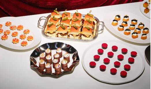 Assorted party snacks on white plates, including mini sandwiches, chocolate candies, red gummies, and sushi rolls, arranged on a table.