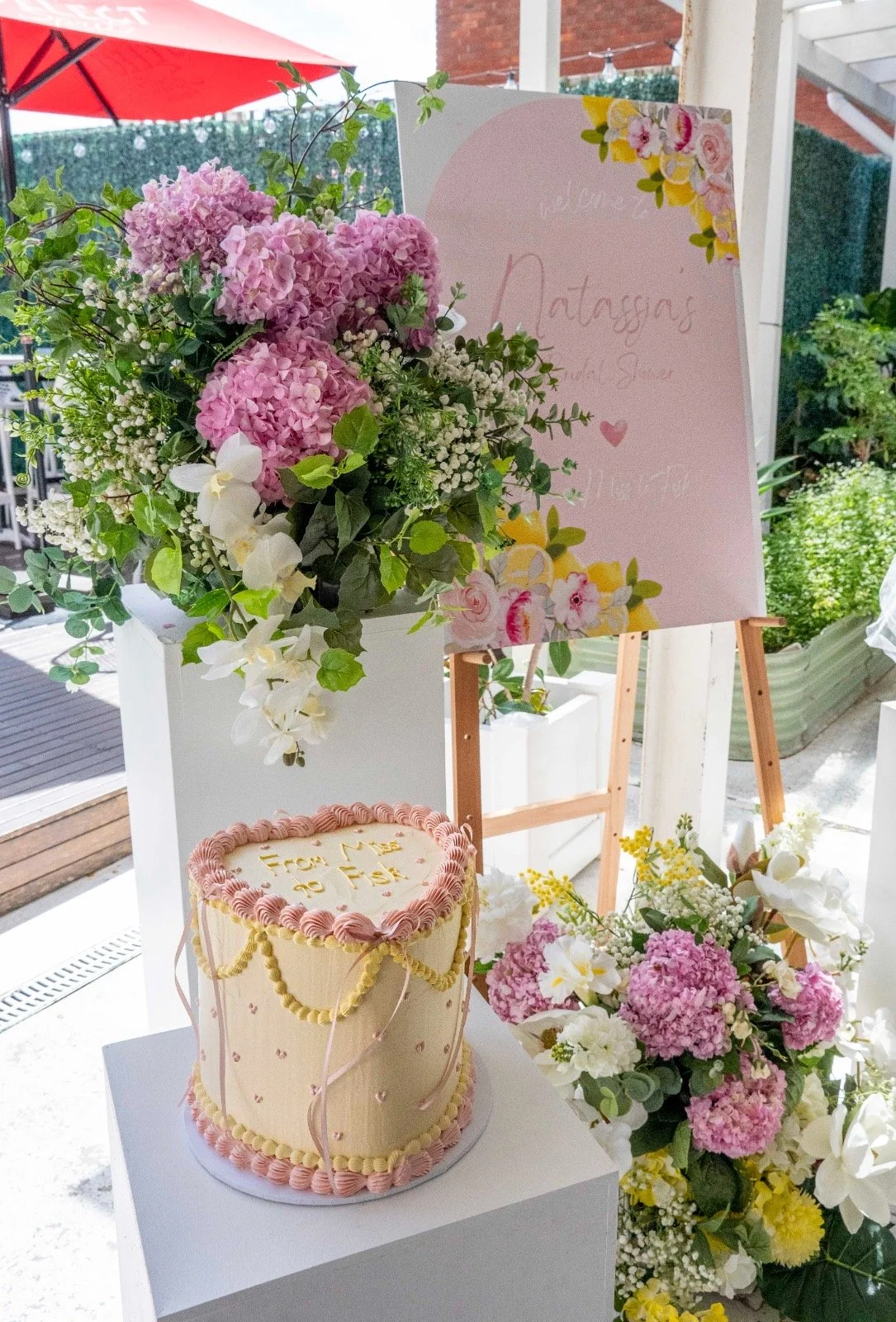 Flower arrangement with pink hydrangeas, white orchids, baby's breath, greenery, a decorated cake with a pink border, and a welcome sign with floral decorations.