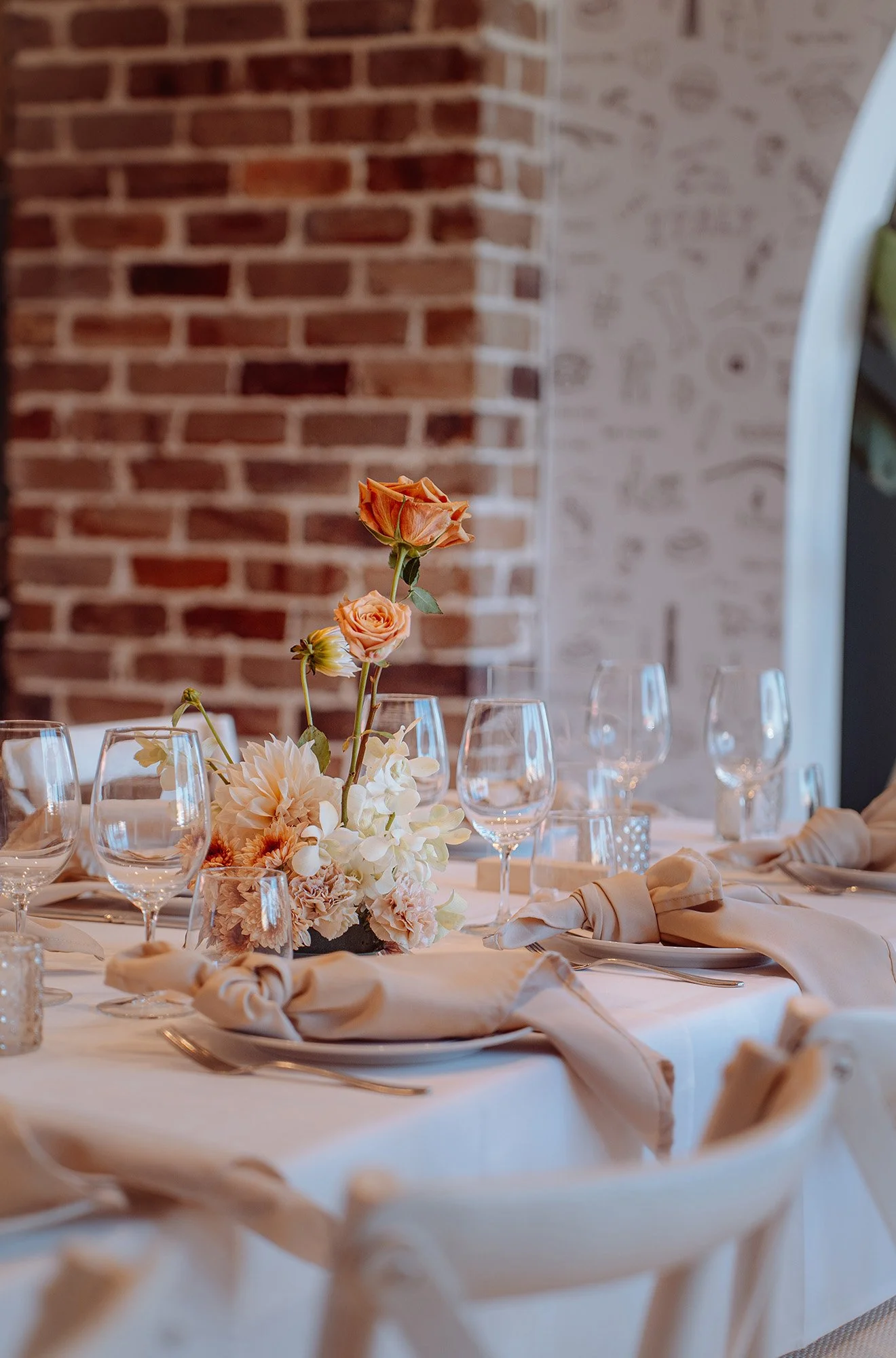 A decorated dining table with a floral centerpiece featuring peach-colored roses and white flowers, set with wine glasses, napkins, and plates in a room with exposed brick walls.