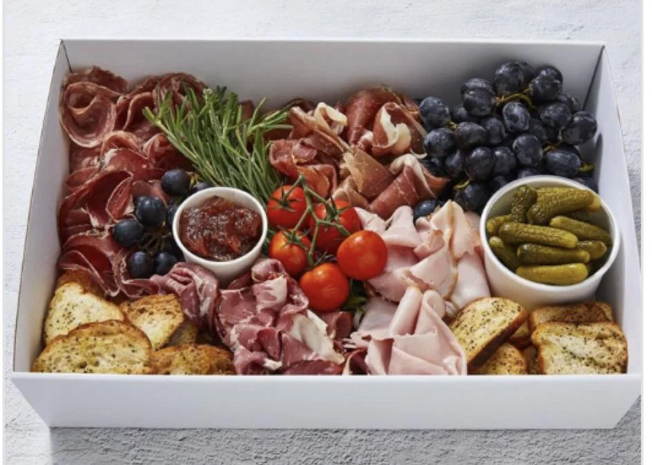 Charcuterie board with sliced meats, cherry tomatoes, blueberries, grapes, pesto, bread slices, and bowls of jam and pickles.